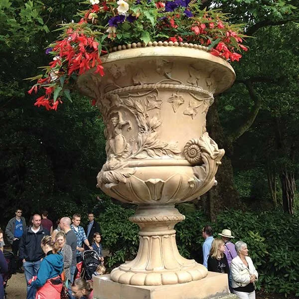 Large decorative stone urn filled with colorful flowers, surrounded by people in a park or garden setting with trees in the background.