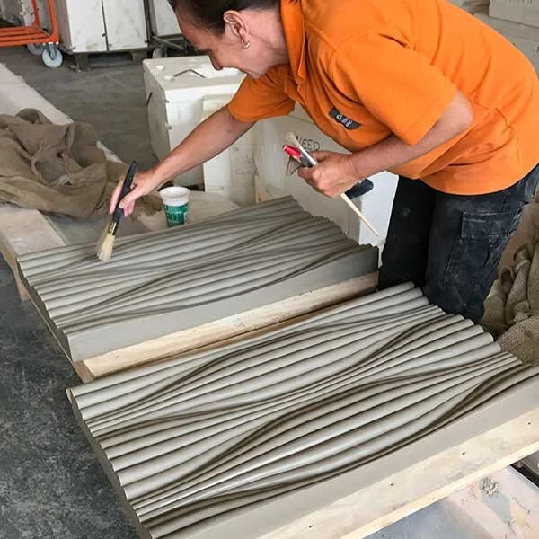 A person wearing an orange shirt is decorating ceramic tiles with a textured wave pattern. They are holding a brush and a tool, working on the surface of the tiles in a workshop setting.