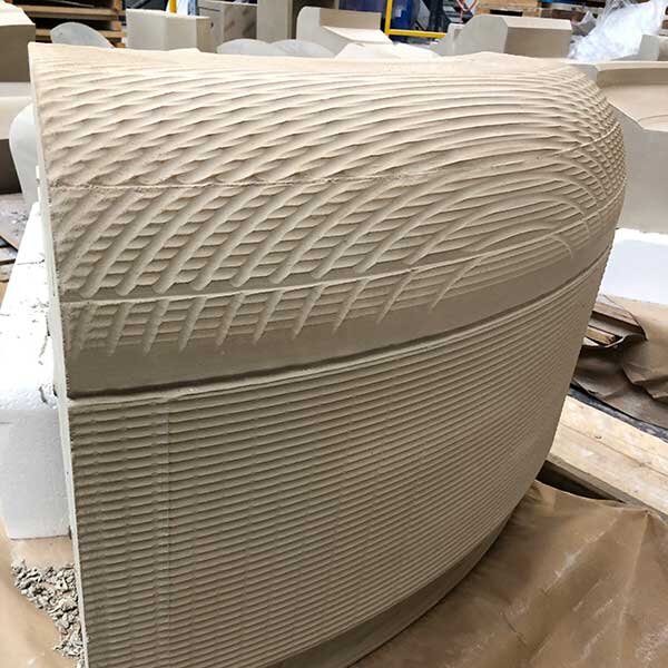 Close-up of a textured beige or light-colored curved surface with a woven or ridged pattern, likely part of a piece of furniture or decorative object in a workshop setting.