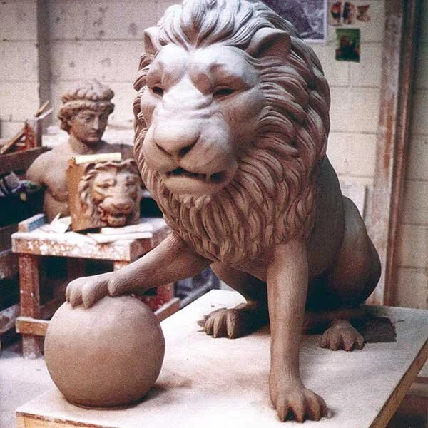 A finished clay sculpture of a lion with a detailed mane, sitting on a workbench in a sculpture studio, with additional sculpture projects and materials visible in the background.
