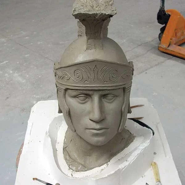 A clay sculpture of a male figure wearing a helmet with detailed carvings, in an artist's studio.