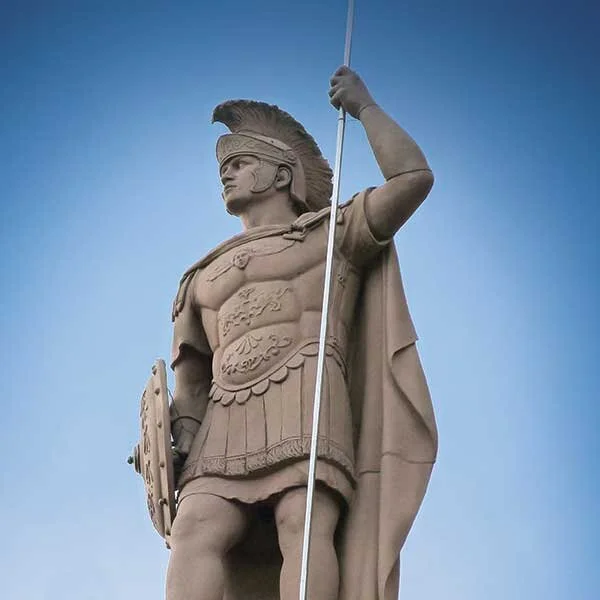 Statue of a Roman soldier holding a spear and a shield, wearing a helmet with a crest, and a cape, against a blue sky.