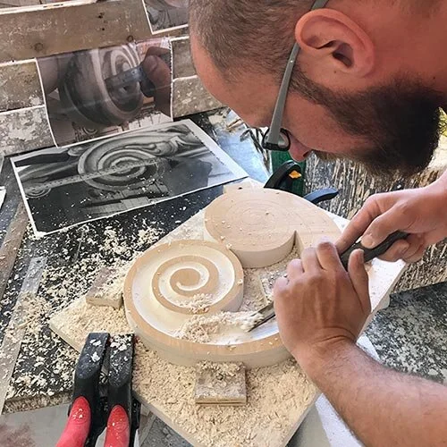A man carving a spiral design into a piece of wood using a chisel and mallet, with woodworking tools and reference images nearby.