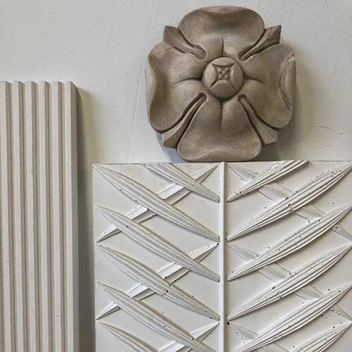 A close-up of an art installation featuring a circular, flower-shaped sculpture mounted on a white wall, with perpendicular white structural elements with diagonal lines below, and a vertically ribbed beige panel to the left.