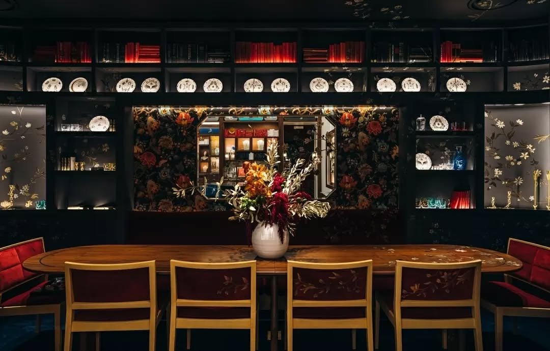 An oblong dining room, wrapped in rich millwork, showcases the homeware collection. A hand-painted floral motif, inspired by the brand's resilience tenet, flows across the ceiling, walls, table, chairs, and extends seamlessly onto the carpet below.

