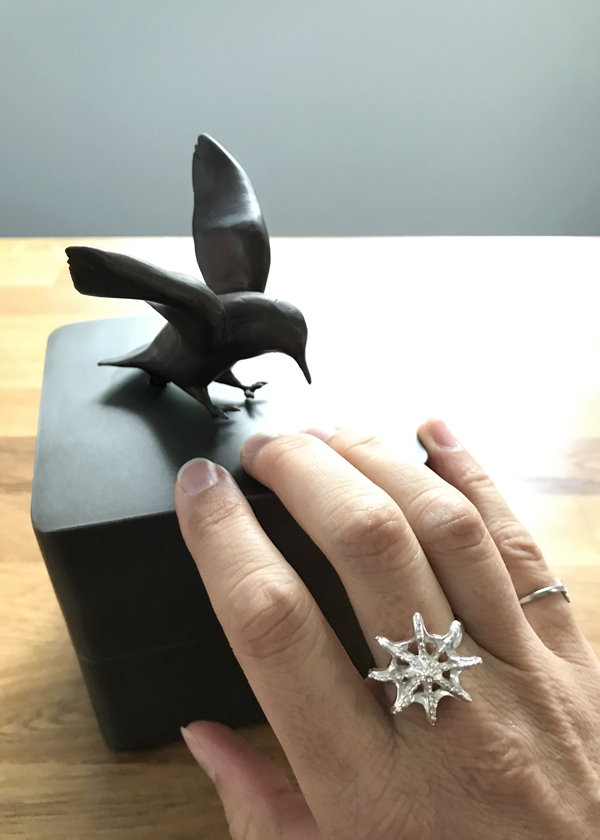 bronze bird in flight on top of steel cube music box sculpture held by hand wearing silver sea star ring. Interior of music box has crystal landscape and fish out of water.