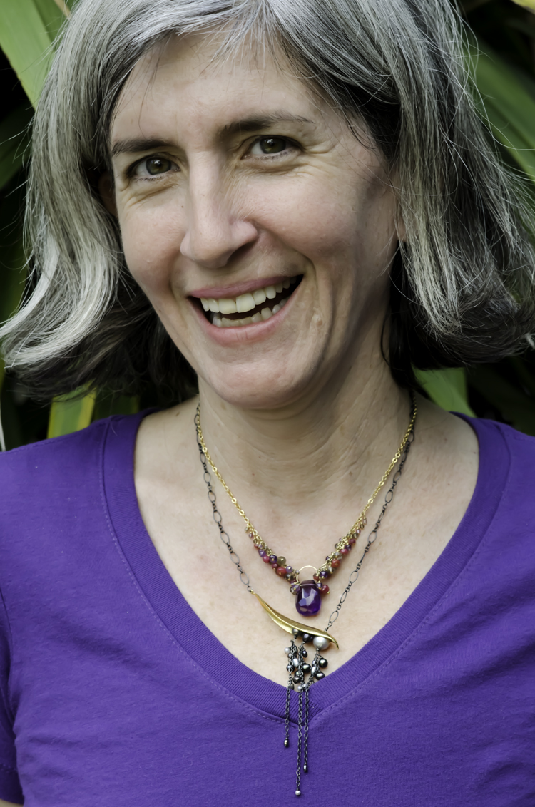 woman with grey hair and style. Wearing purple t-shirt, gold, purple, and red gemstone necklace layered with gold, black silver, and grey pearl asymmetrical necklace