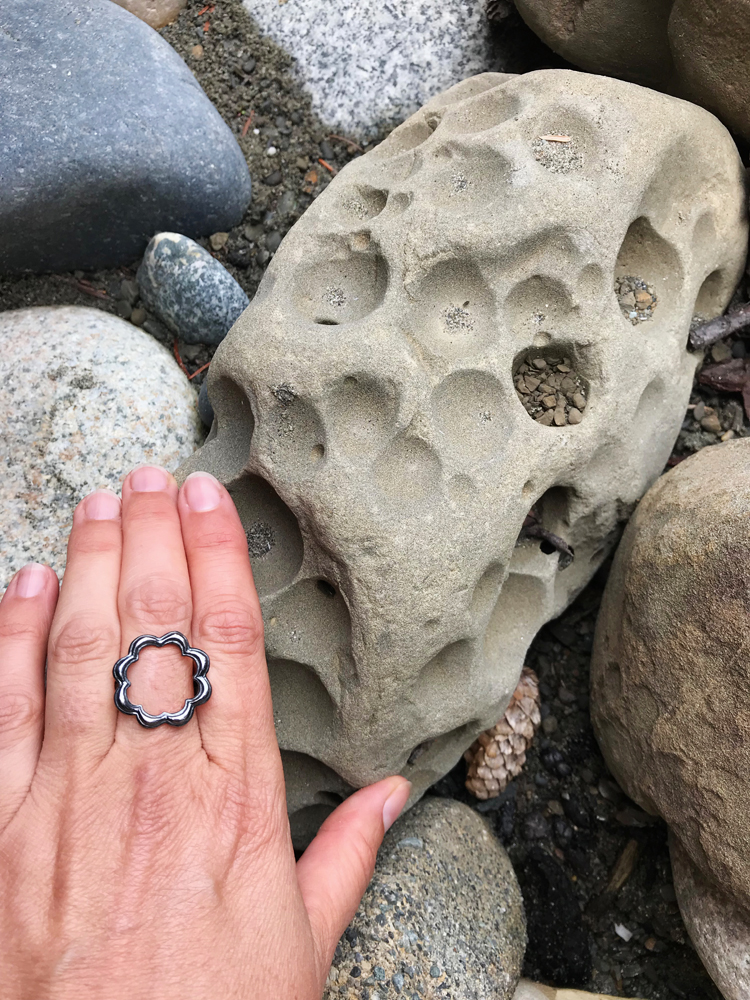 hand wearing black sterling silver scallop circle ring next to grey rock with circle holes. Blue, grey, and white speckled rocks. Shi Shi Beach Washington adventure