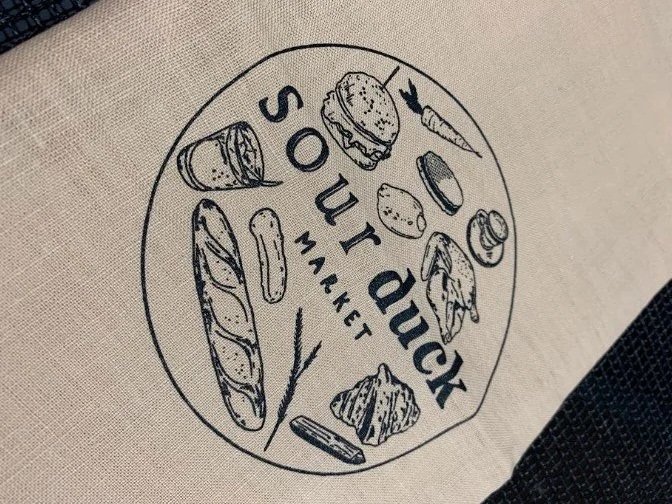Tote bag featuring "Sour Duck Market" and illustrations of food.