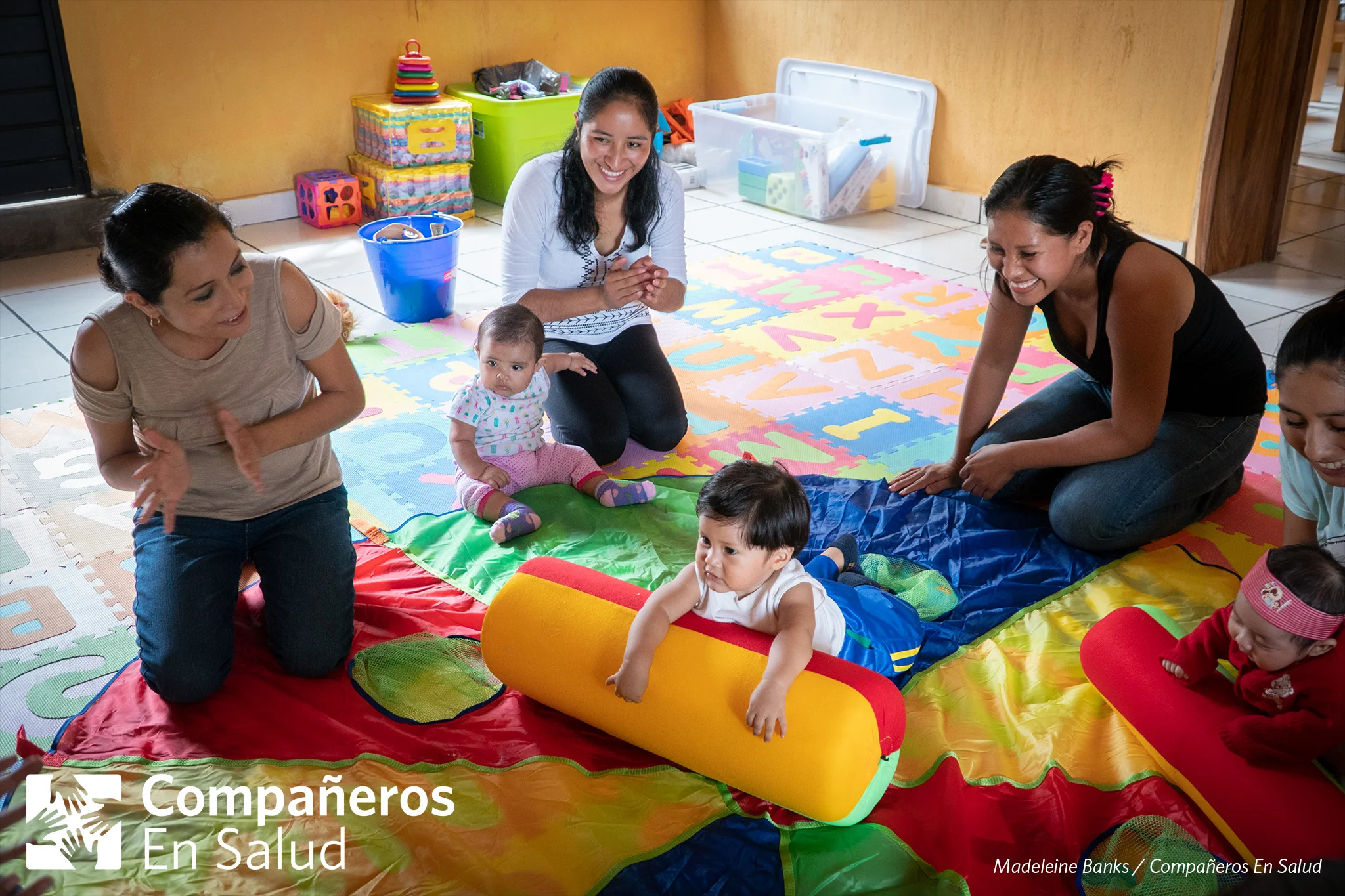 La maestra Luisa Fernanda Estrella Maza y el grupo de madres en el curso de desarrollo infantil y estimulación temprana animan al niño Darek mientras él trata de cruzar el tapete.