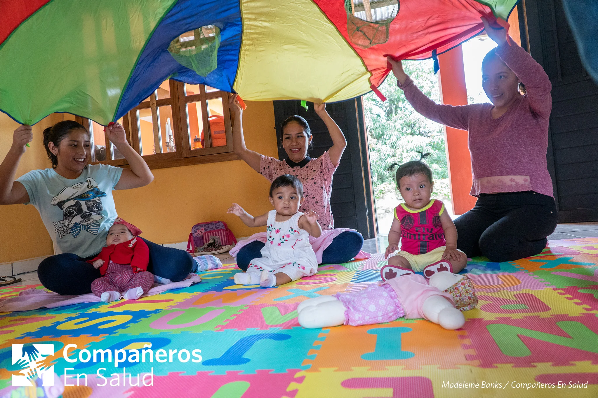 Mamas de Jaltenango de la Paz agitan un paracaídas sobre sus hijas y cantan sobre los colores durante un curso de desarrollo infantil y estimulación temprana.