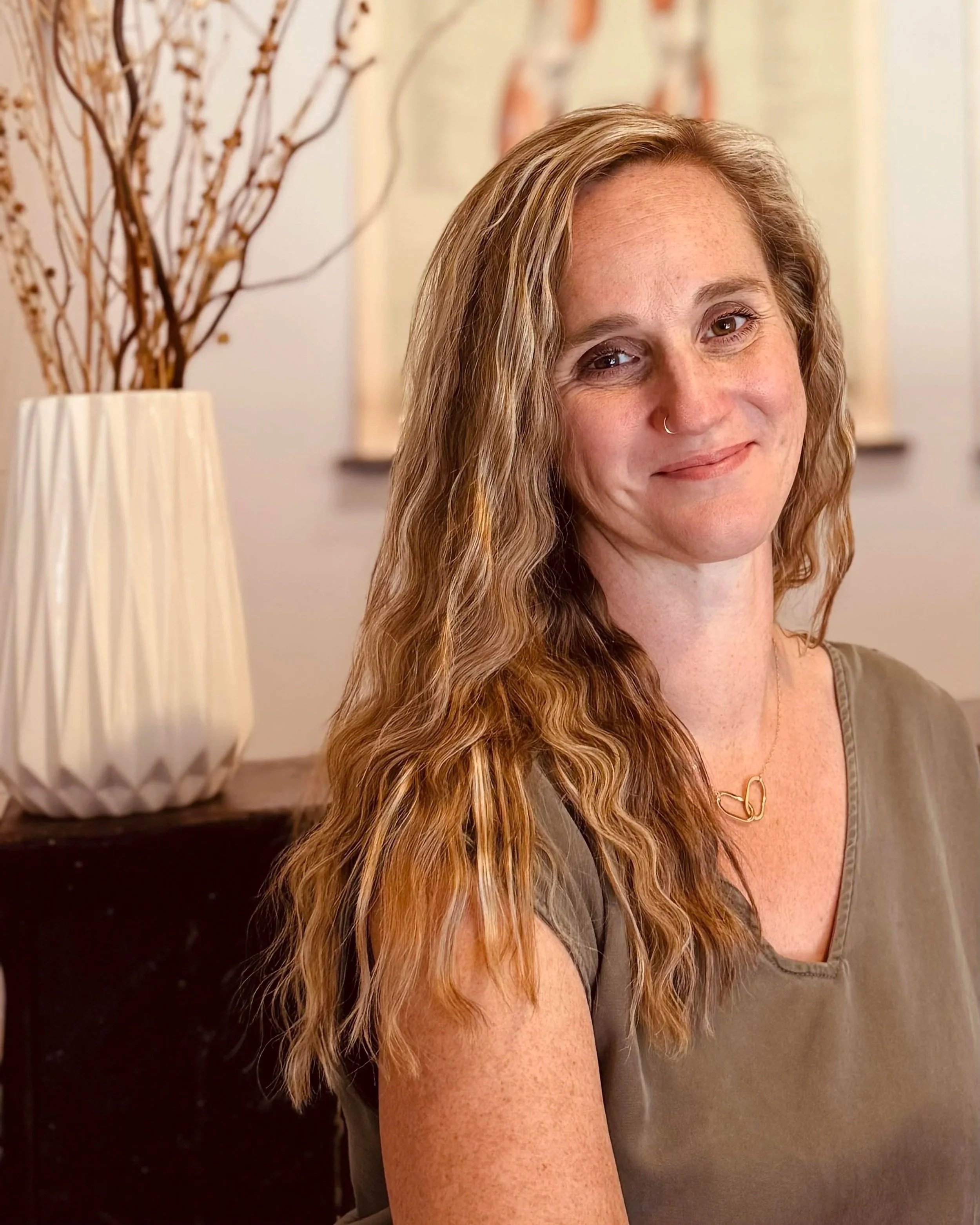 A woman with long, wavy hair and a nose piercing smiling, wearing a gray sleeveless top and a gold heart-shaped necklace, standing indoors with a vase of dried branches and a medical poster in the background.