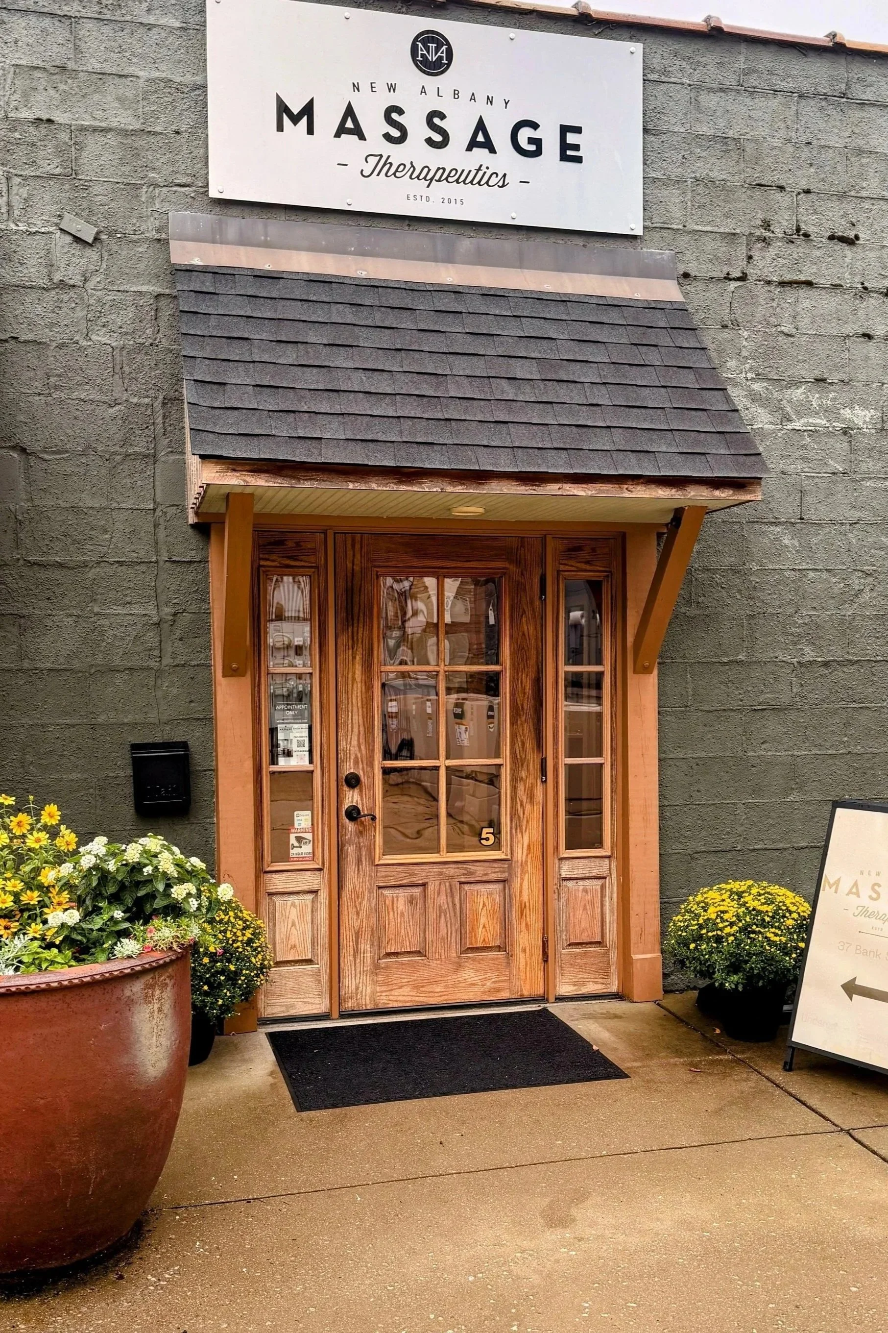 The entrance to a massage therapy business with a wooden door, potted plants with flowers on either side, and a sign above that reads 'New Albany Massage Therapuetics'. There is also an A-frame sign on the right side with the same business name and an arrow pointing left.