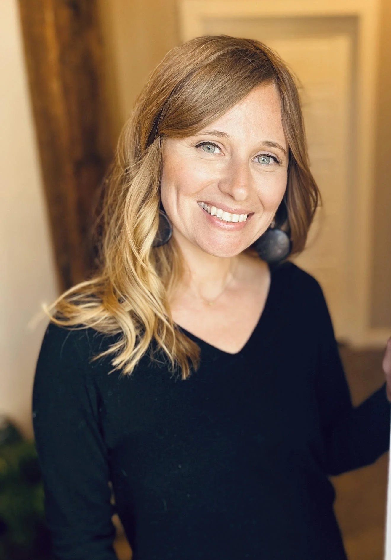 A woman with long, wavy blonde hair and blue eyes smiling at the camera, wearing a black top and large earrings in an indoor setting.