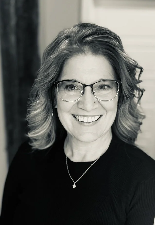 A smiling woman with wavy hair, glasses, and a necklace.