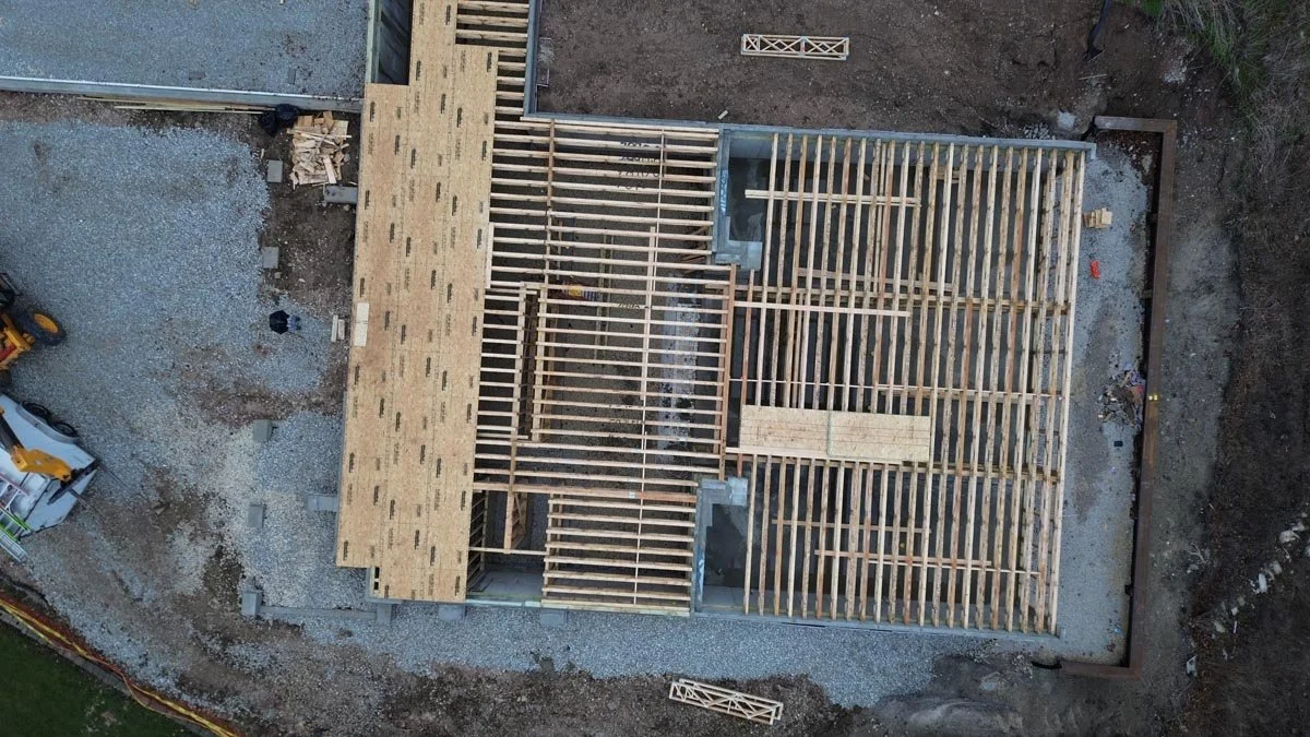  Overhead aerial view of floor framing partly covered in plywood subfloor, next to a gravel yard and equipment. 
