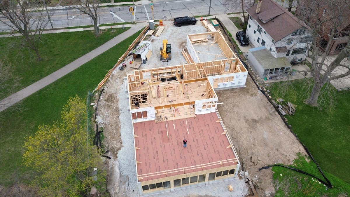  Aerial view of first-floor wall framing and a red-sheathed lower-level deck, with a worker on site and the street behind. 