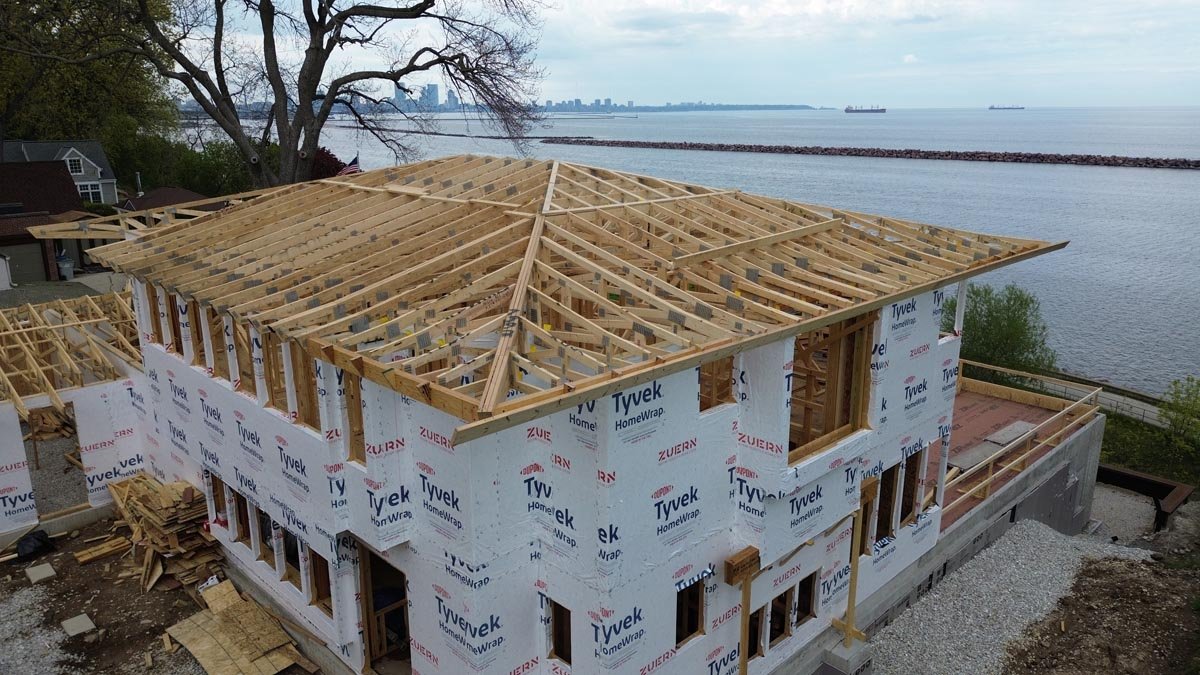  Aerial view of a house-wrapped home with a completed hip roof frame, overlooking Lake Michigan, a breakwater and a city skyline. 