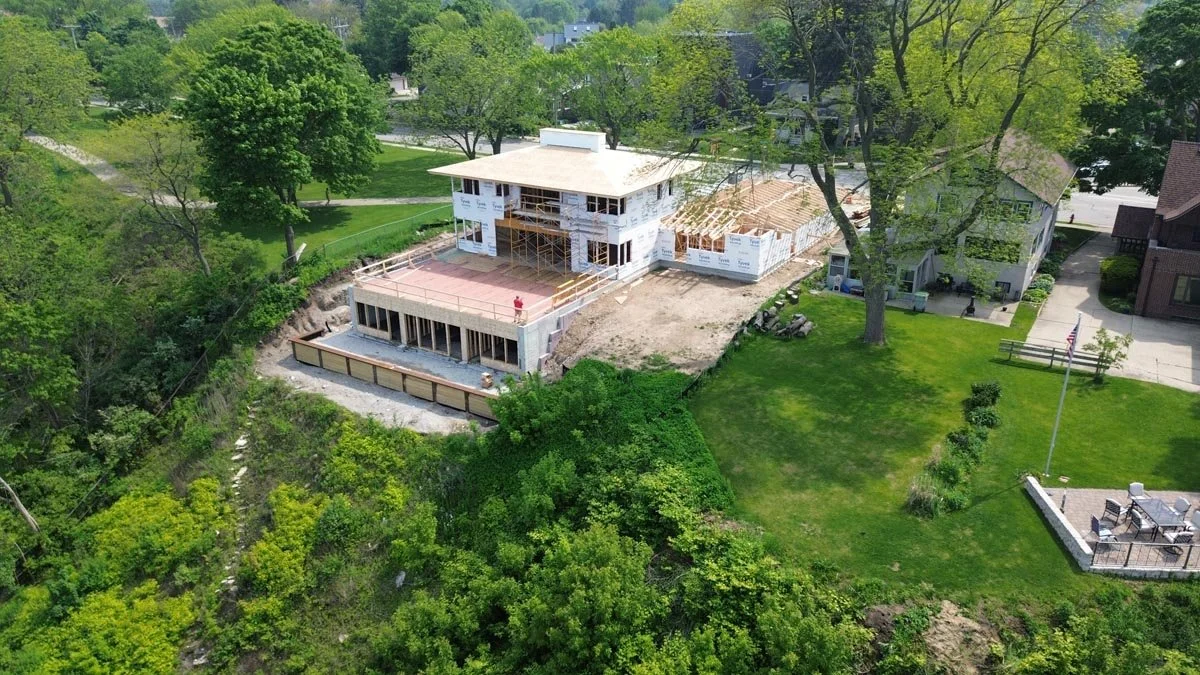   Aerial view from the north of the house under construction on a wooded bluff, with a neighboring yard and flagpole to the right. 