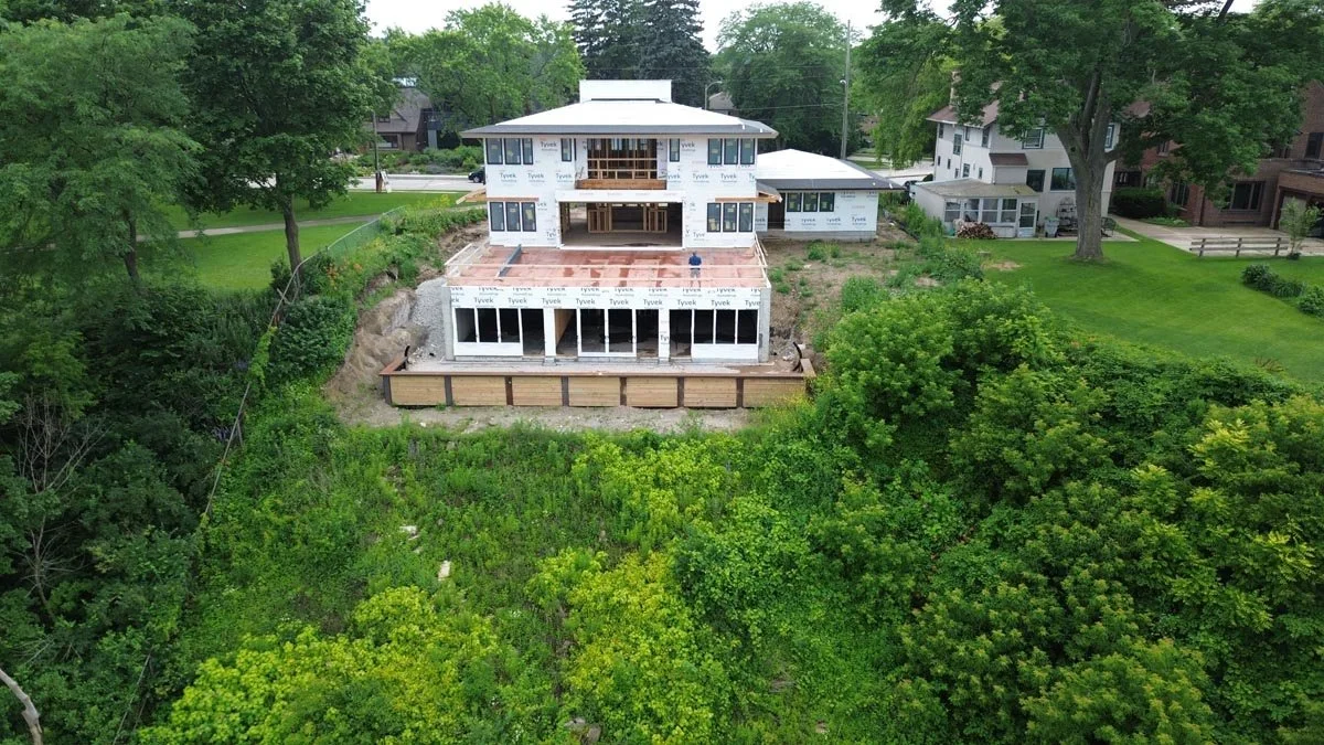  Aerial view from the lake of the wrapped two-story house with large window openings, set above a steep vegetated bluff. 