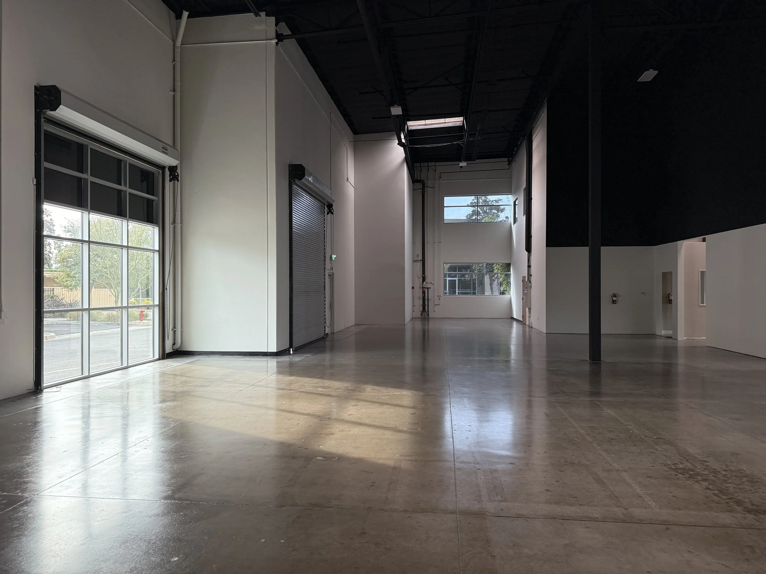 Empty commercial space with polished concrete floors, large windows, a rolled metal door, and high ceilings with exposed ductwork and a black ceiling section.