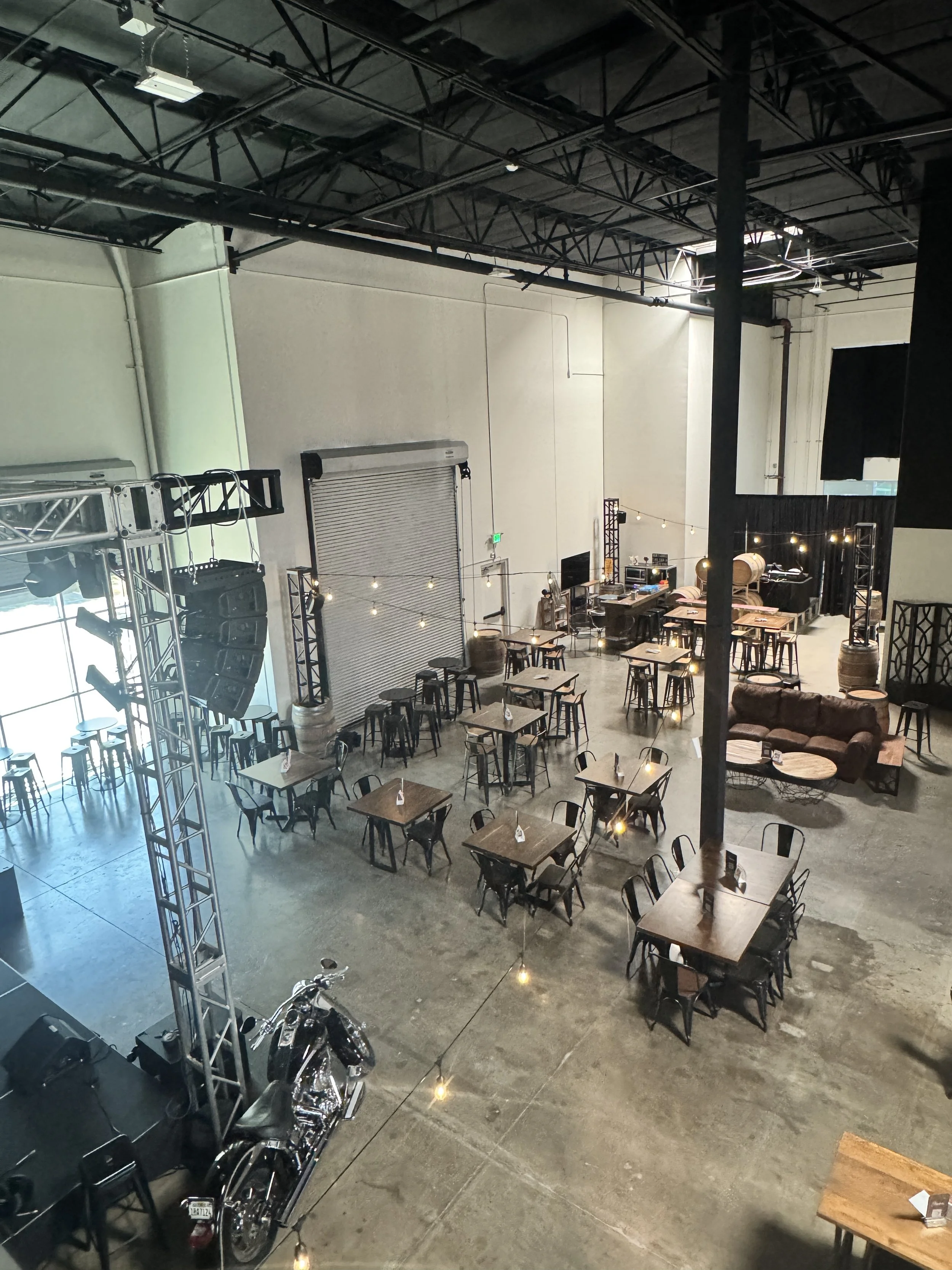 Interior of a modern event space or brewery with tables and chairs, a bar area, and motorcycles parked near the entrance. The space features high ceilings, industrial lighting, and string lights creating a cozy atmosphere.
