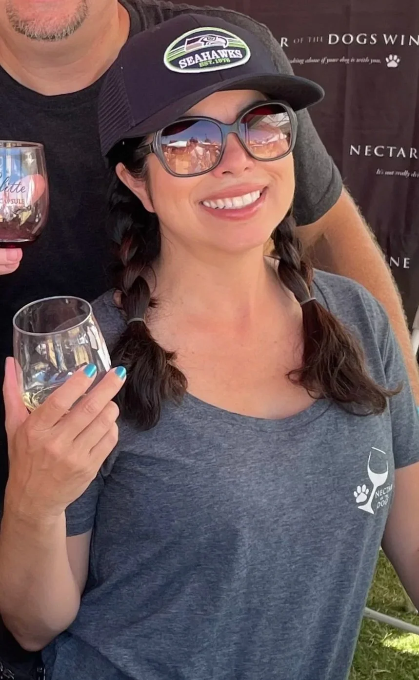 A woman with braided hair wearing sunglasses, a Seattle Seahawks cap, and a gray t-shirt holding a glass of white wine. She is smiling at the camera.
