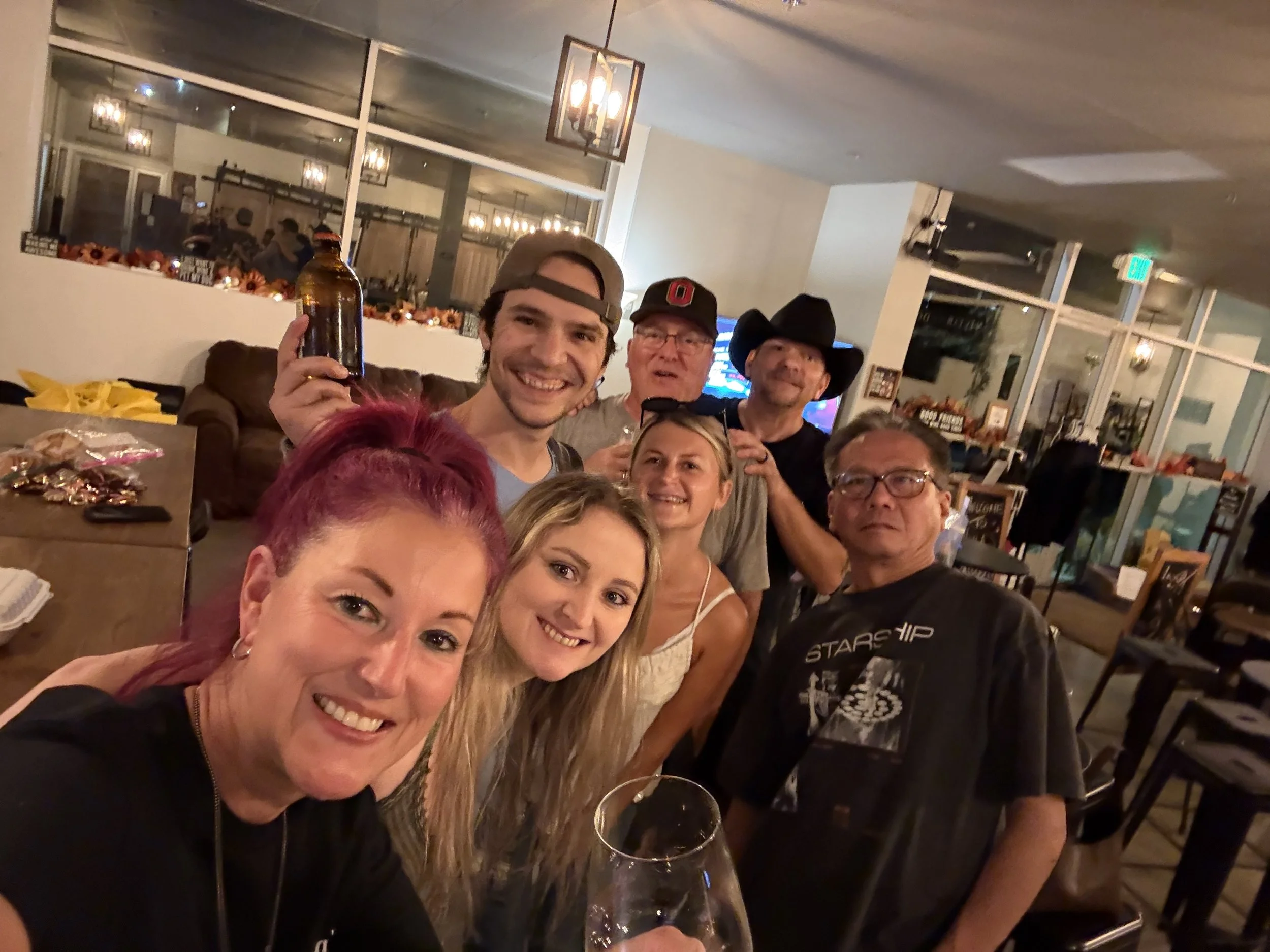 A group of seven people smiling at the camera in a warmly lit restaurant or bar, with some holding drinks, including a man in a cowboy hat, a woman with pink hair, and a man wearing glasses.