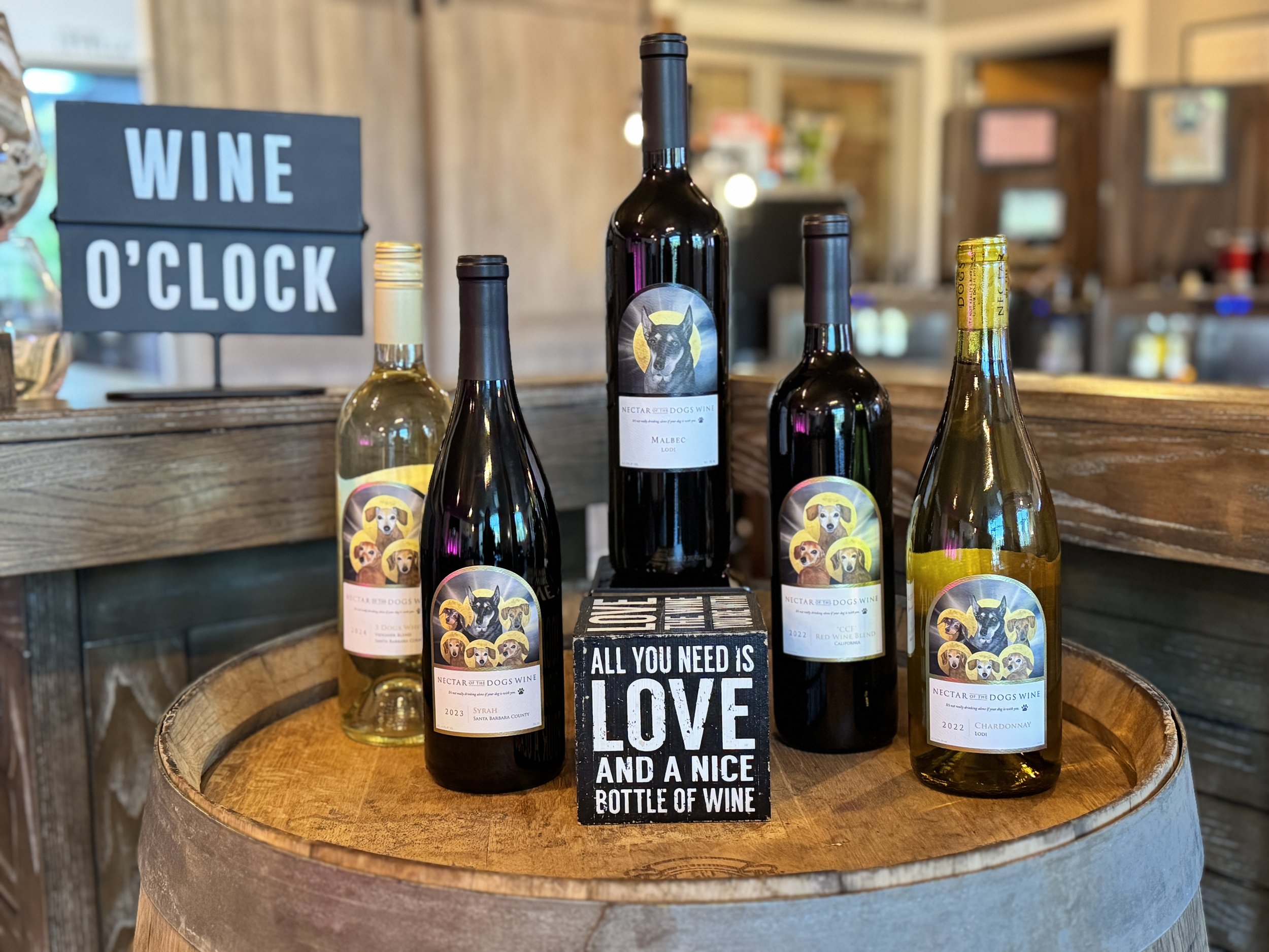 Several bottles of wine on a wooden barrel with a sign reading "WINE O'CLOCK" behind them. There is also a black and white sign in front of the bottles that says "All you need is love and a nice bottle of wine."