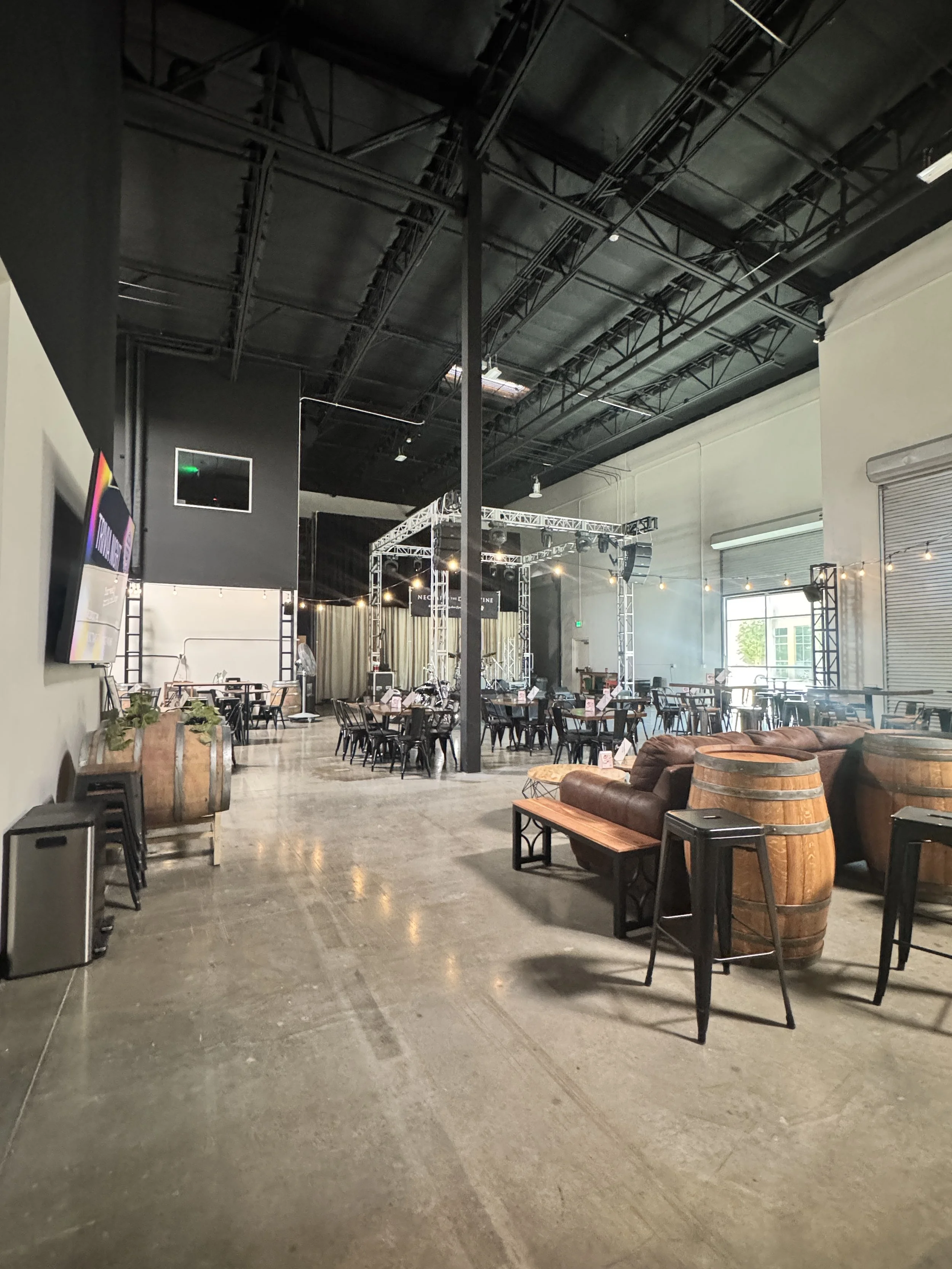 A spacious, industrial-style event venue with high ceilings, black metal beams, and large windows. The room is decorated with string lights and has a mix of chairs, tables, and lounge seating, including leather armchairs and wine barrels. There is a stage setup with scaffolding and lighting equipment.