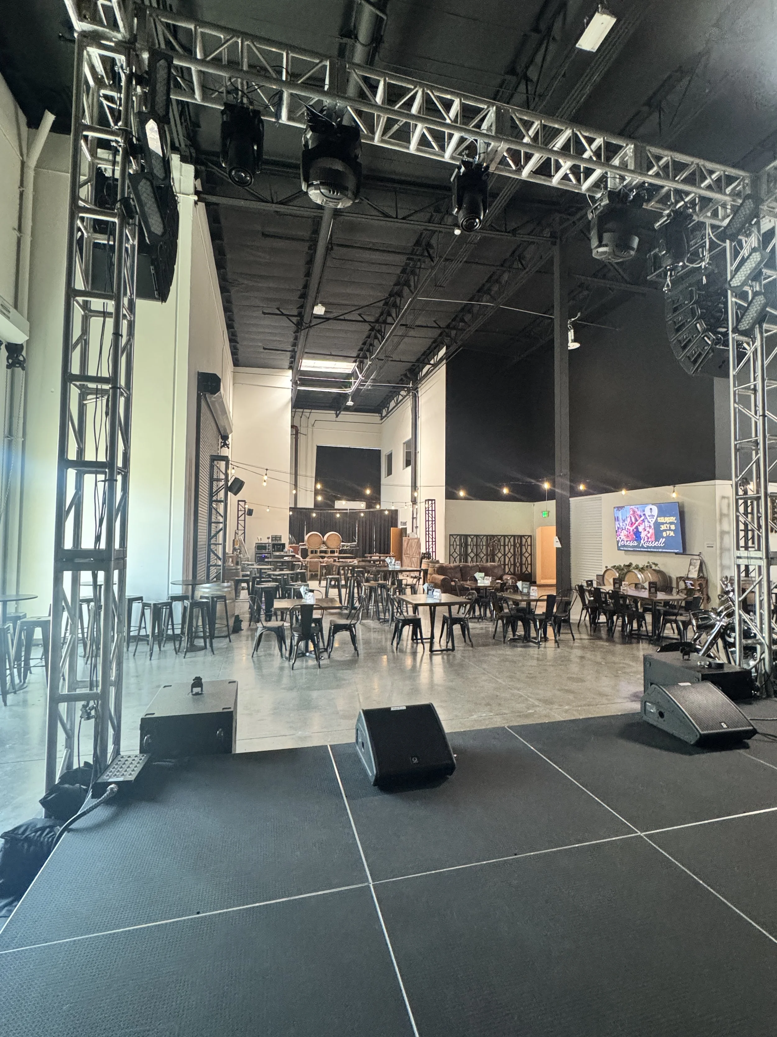 Stage with black floor and black curtains, musical instruments, and an arrangement of chairs and tables in a room with high ceiling and lighting equipment.