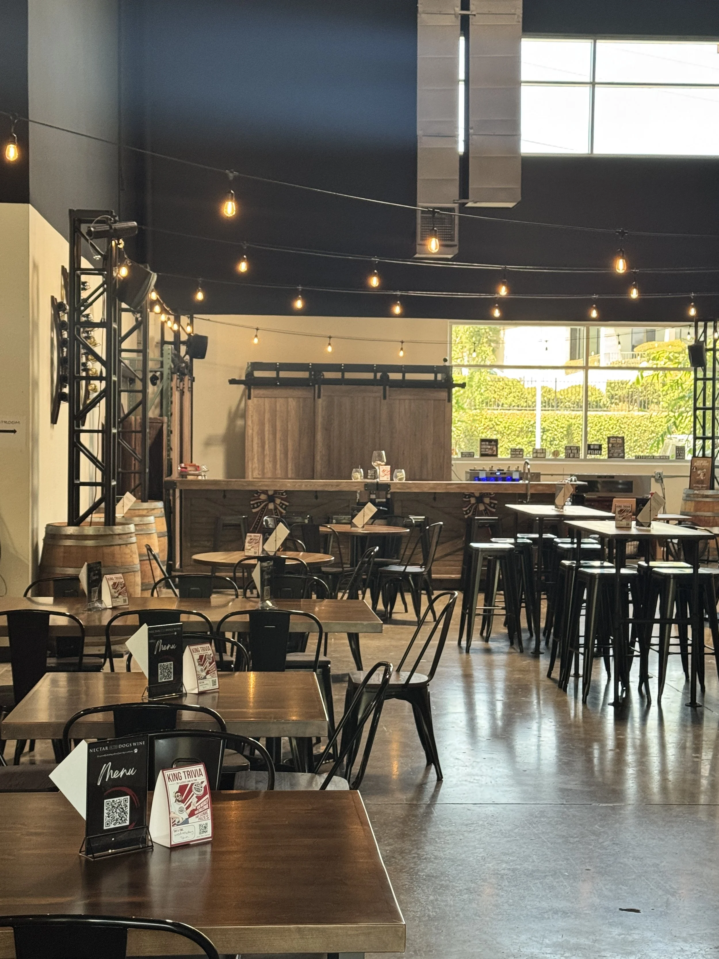 Interior of a modern restaurant or cafe with wooden tables, black chairs, menu stands, and string lights hanging from the ceiling. Large windows let in natural light, showing a view of trees outside.