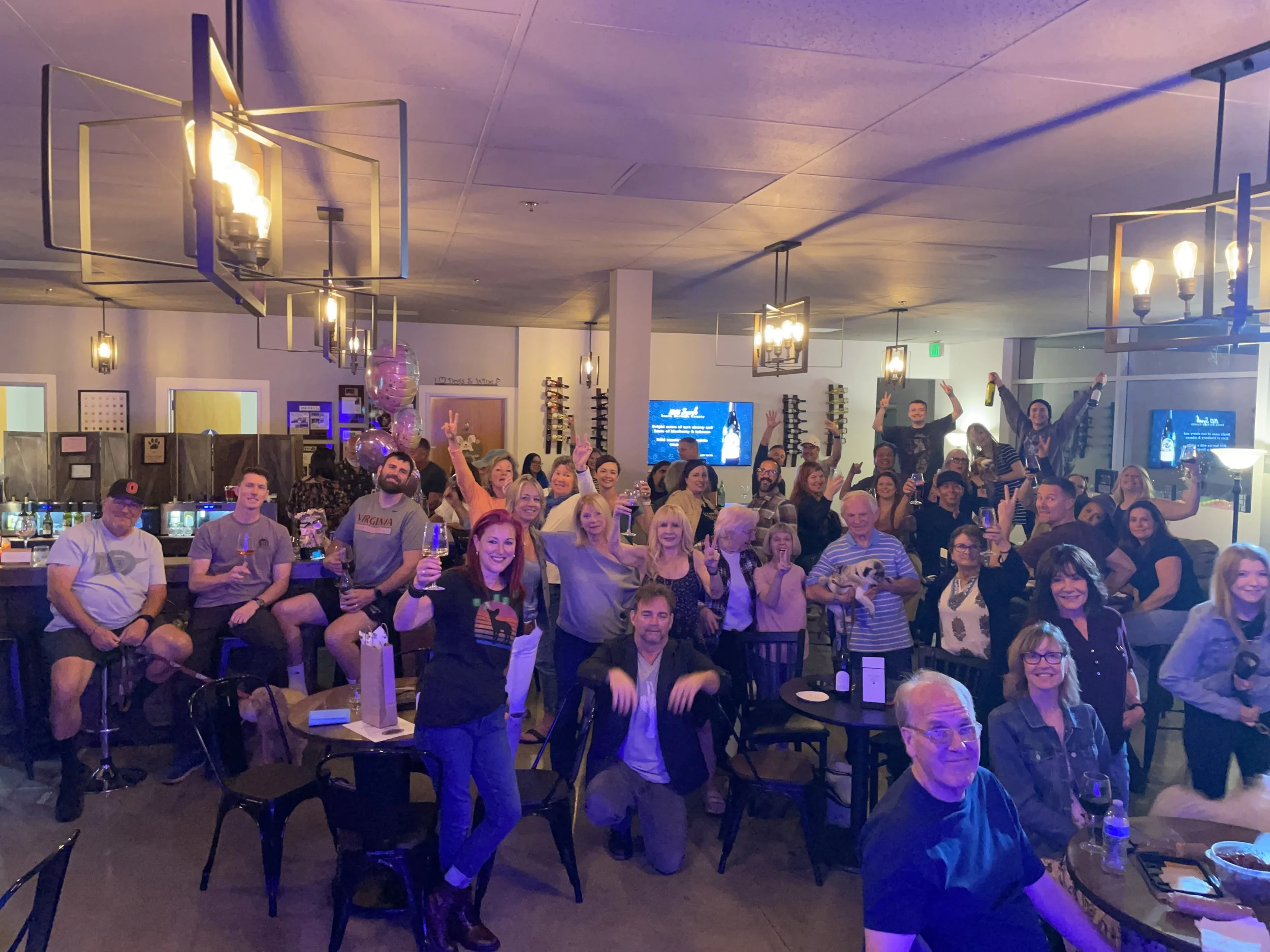 A large group of people gathered in a wine tasting room, smiling and holding drinks, celebrating together. The space is decorated with modern lighting and has a lively atmosphere.