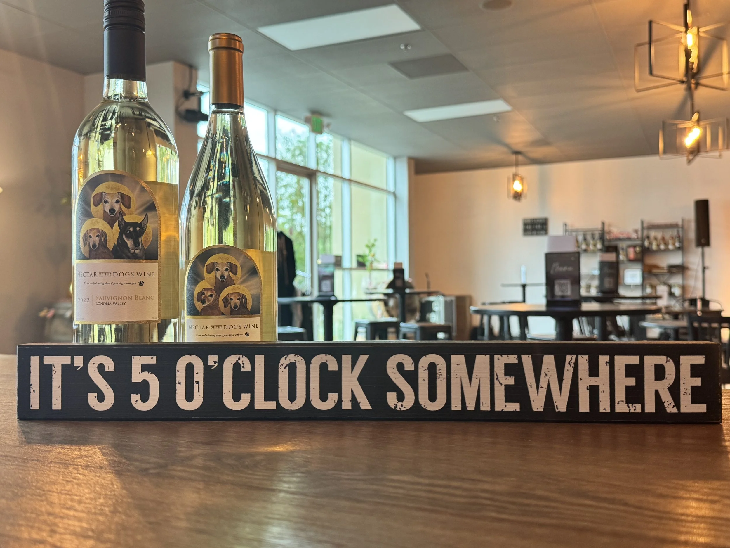 Two bottles of wine labeled 'Nectar of the Dogs Wine' on a table with a sign that reads 'It's 5 o'clock somewhere.' In the background, a spacious, well-lit room with large windows, tables, and hanging light fixtures.