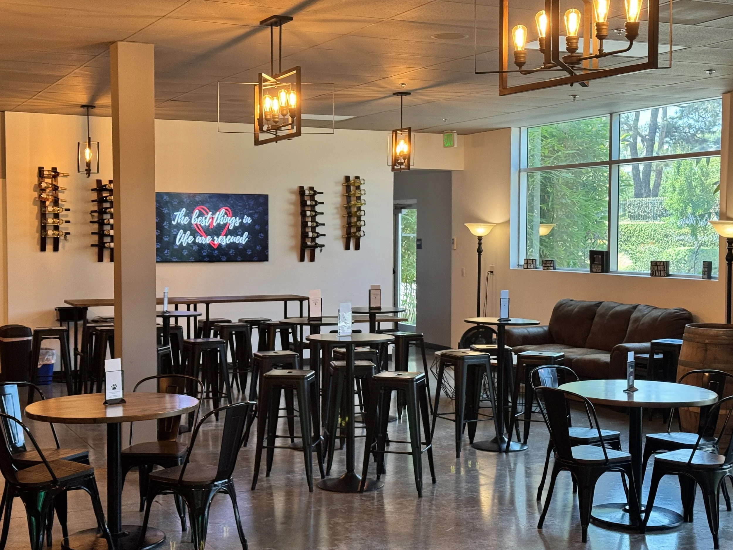 Interior of a wine tasting room with round tables and black chairs, a leather sofa, wall-mounted wine racks, a wall-mounted TV, and large windows with greenery outside, illuminated by warm hanging and standing lights.