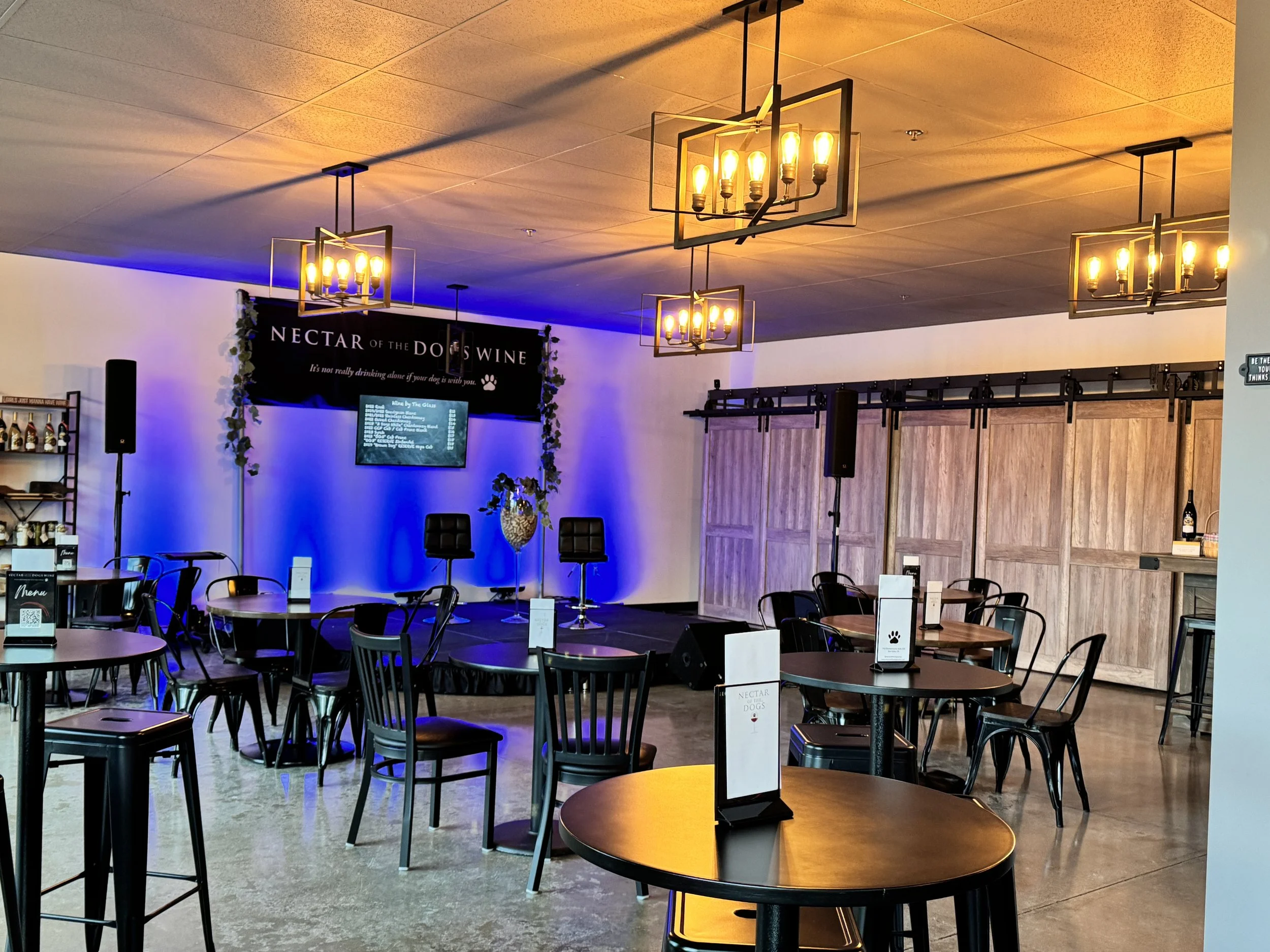 Indoor event space with a small stage, hanging chandeliers, tables and chairs, a menu stand, and a backdrop with the text 'NECTAR OF THE DOG'S WINE,' decorated with vine garlands.