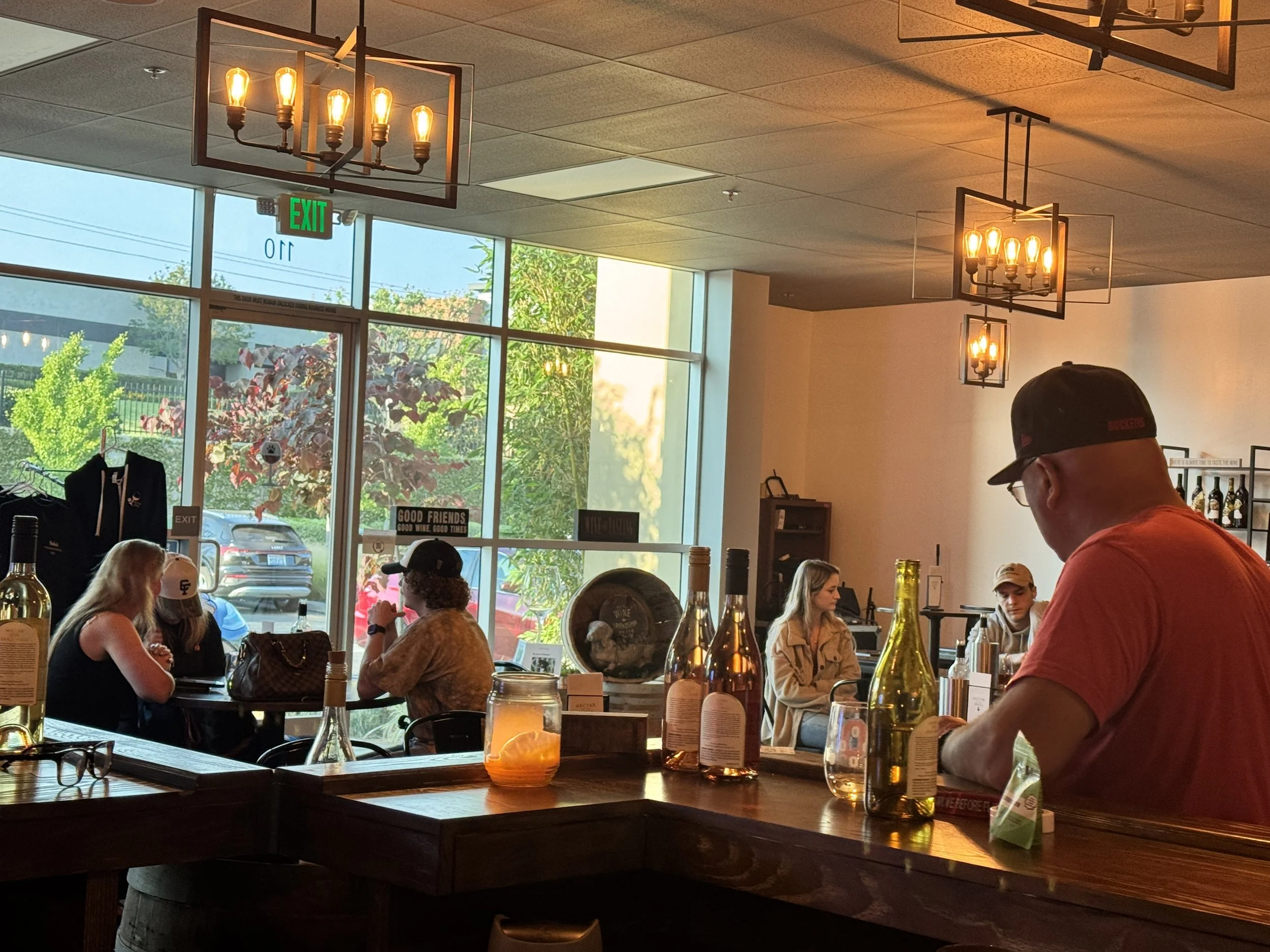 Inside a wine tasting room with large windows showing trees and parking lot outside. Several patrons seated at tables and bar, with bottles and drinks visible. Warm lighting and modern decor.