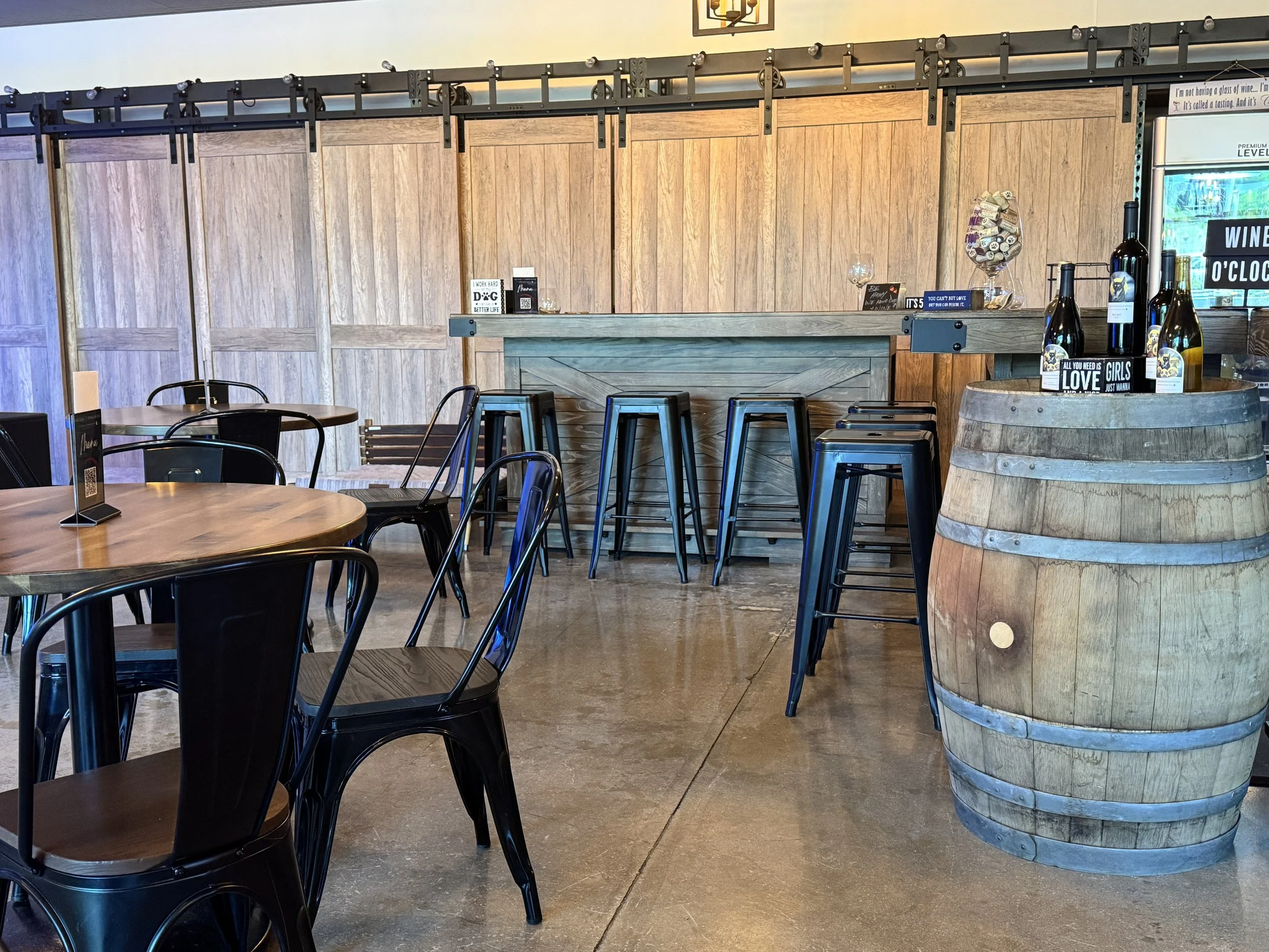 Wine tasting room with wooden tables, black metal chairs, a wooden bar counter with black bar stools, and a large wooden barrel used as a table with bottles of wine on top.