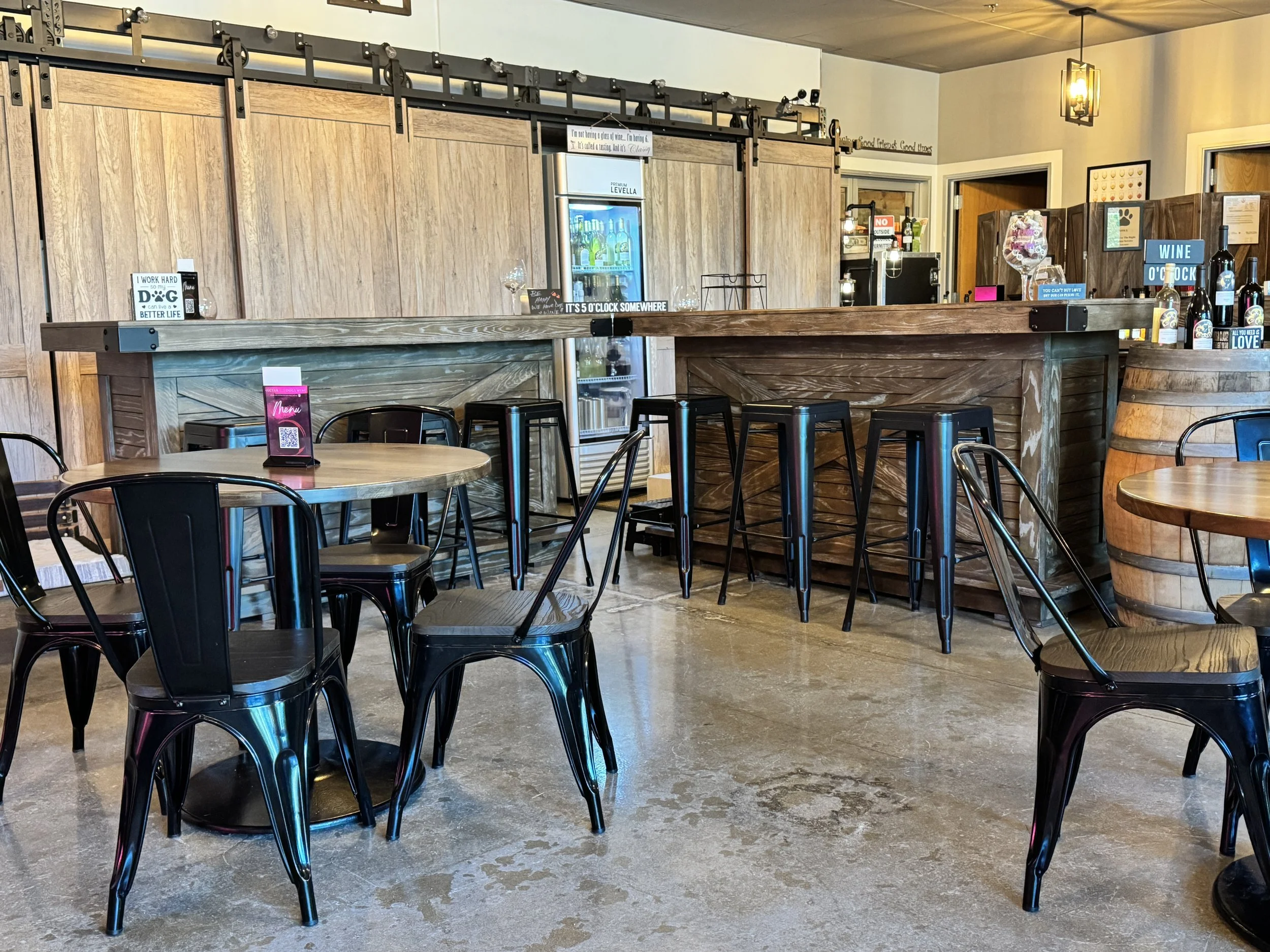 Interior of a wine tasting room with wooden furniture, black metal chairs, a wooden counter, and various signs and bottles on display.