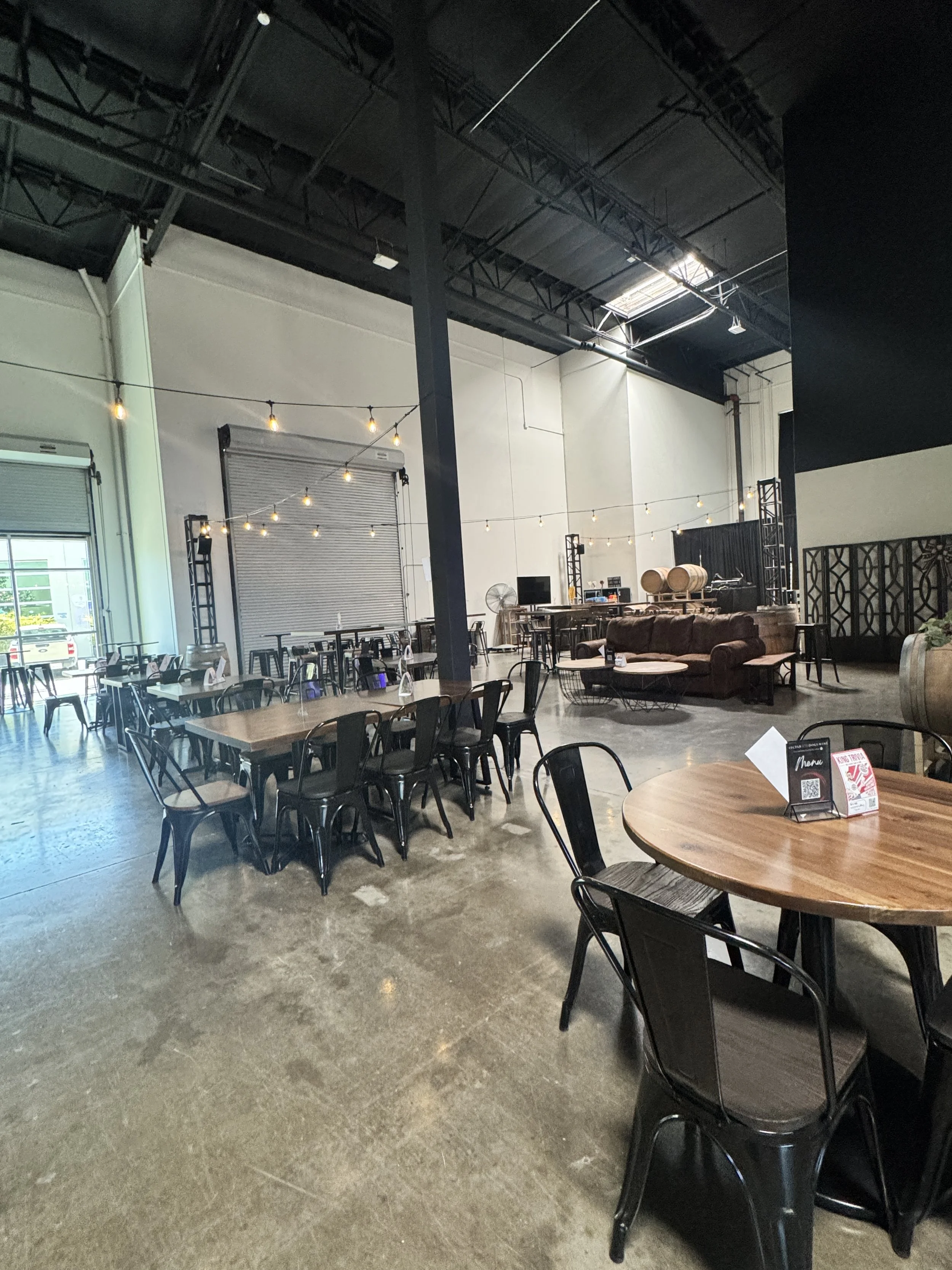 Interior of a modern restaurant or cafe with wooden tables and black chairs, a brown couch, string lights hanging from the ceiling, and a large open space with a high ceiling and industrial style decor.