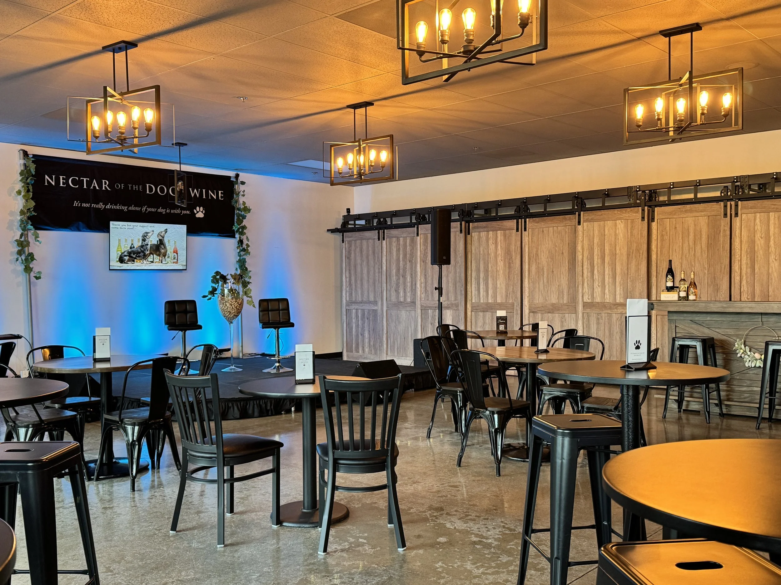 An empty indoor venue with round tables and black chairs, a small stage with two black chairs, a large banner reading 'Nectar of the Dog Wine' with a dog paw print, and a mounted television screen. The room features hanging light fixtures and a woode