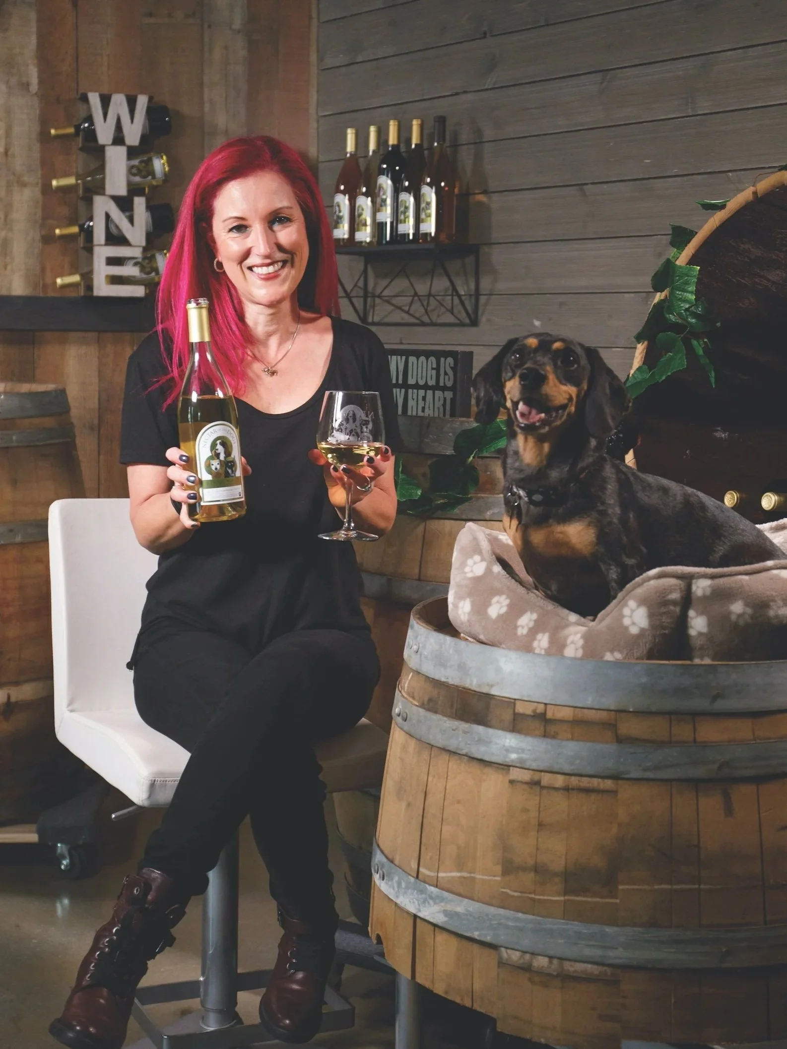 A woman with bright red hair and a black shirt is sitting on a white chair, holding a bottle of wine and a glass of white wine. A small, happy dog is sitting inside a wooden barrel lined with a soft blanket. The background features wine bottles on a shelf and a wooden wall decorated with a large 'WINE' sign and some greenery.