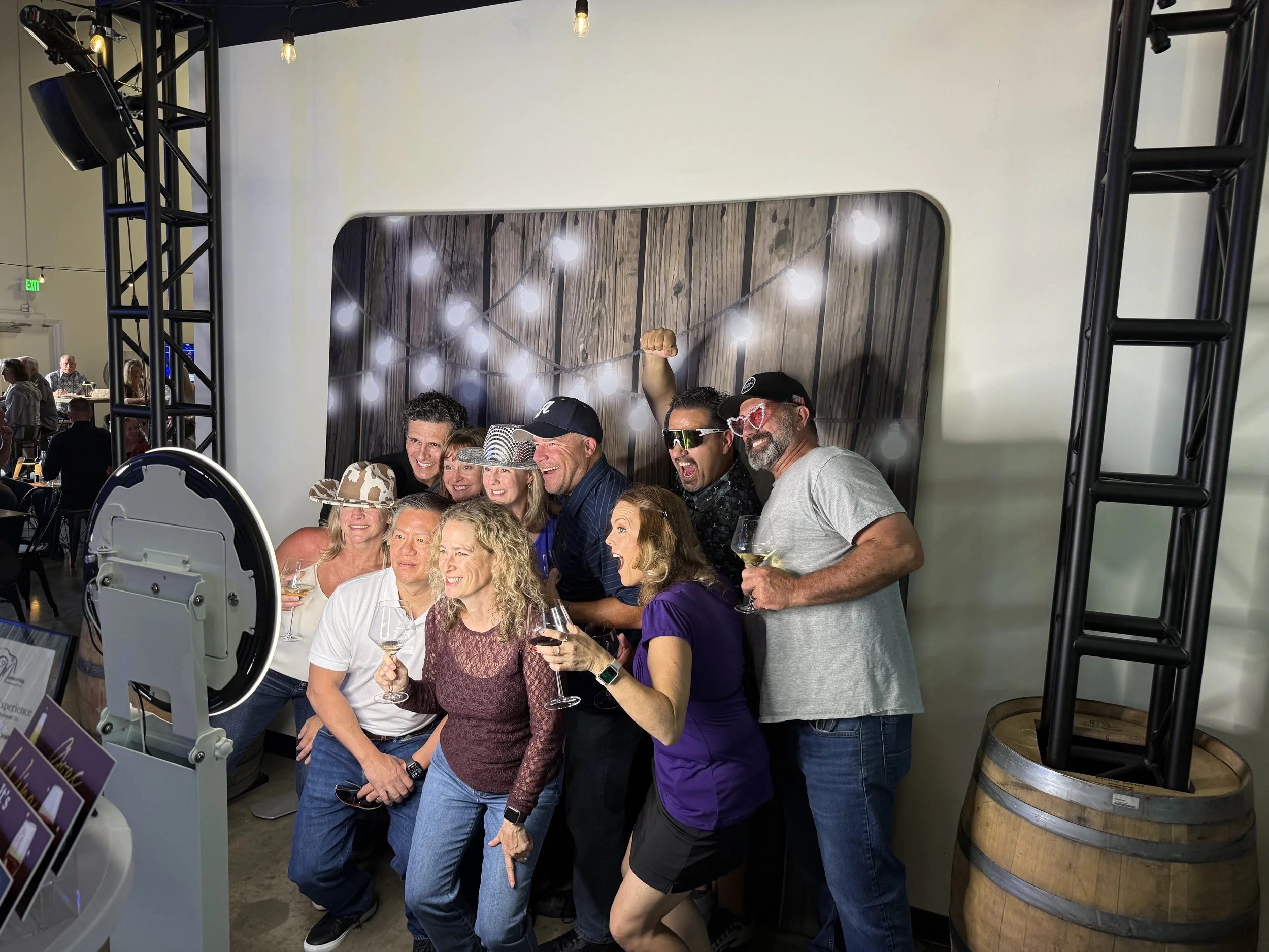 A group of people at a social gathering, smiling and holding wine glasses, posing for a photo indoors with decorative lighting and a wood-themed background.