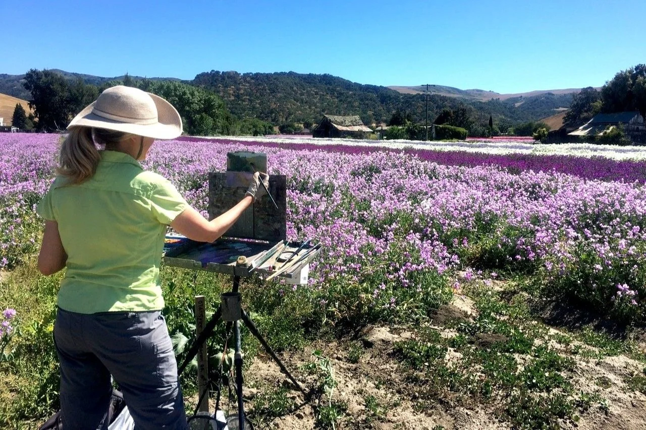 A woman painting on an easel in a colorful flower field with mountains in the background.