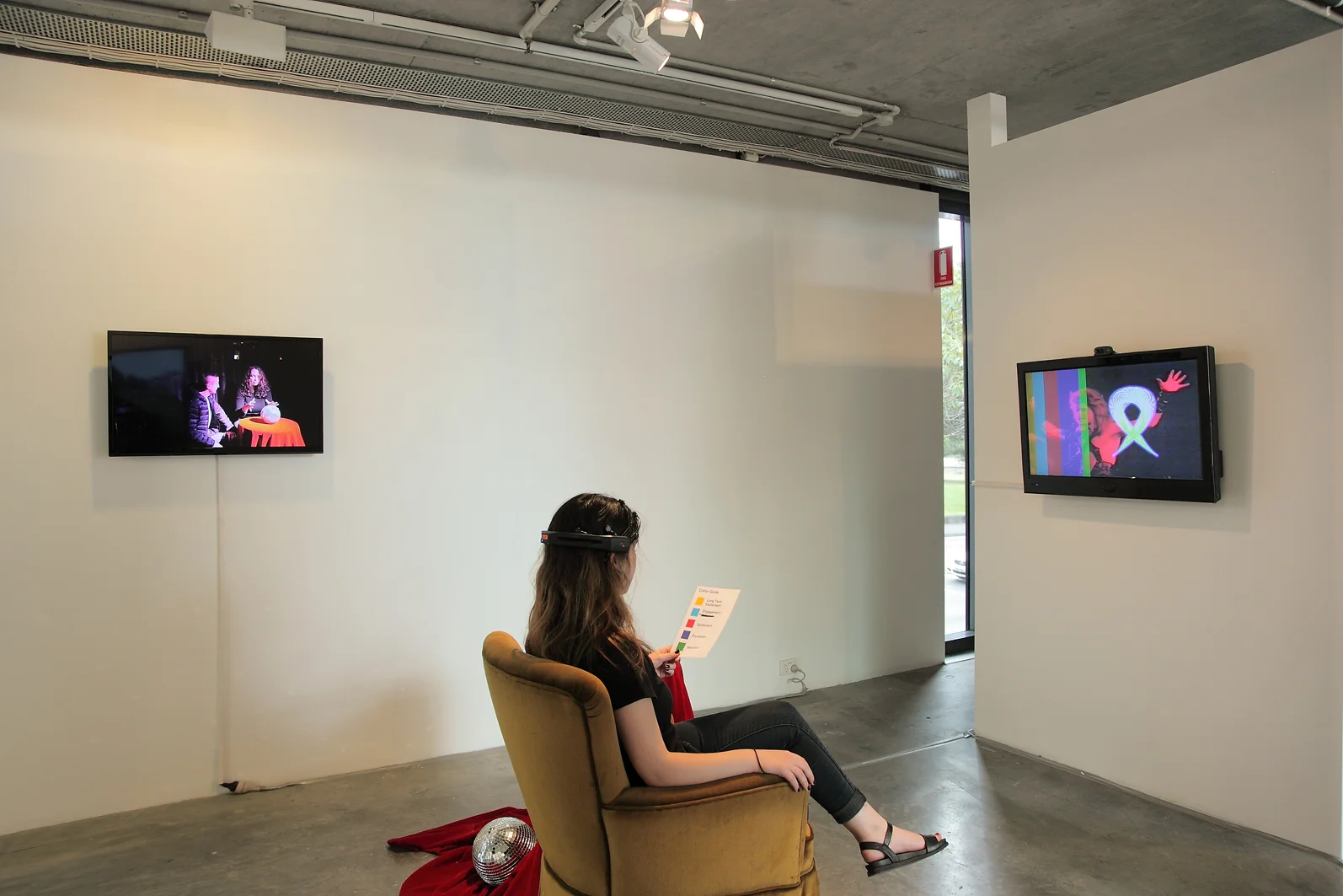 Jacquelene Drinkall, Visual Telepathies exhibition installation view at Verge Gallery, 2015, showing two artworks. LEFT/CENTRE: Jacquelene Drinkall, ‘Disco Ball Gaze’ HD video on HD monitors (colour, audio) disco ball, velvet cloth over small table, documentation of participatory endurance performance at The Cube, Oxford Art Factory, 2014 (no audio was exhibited in the Verge Gallery installation); and Jacquelene Drinkall in collaboration with Warren Armstrong, RIGHT/CENTRE: ‘Hypnosis in Video for Brain Computer Interface’ velvet chair, EEG neuroheadset and participant interaction, live video camera feed, laminated paper map of mental states correlating to colours visualising brain data, 2015 (my video montage of scenes of hypnosis in video, titled ‘Hypnosis in Video’, colour, audio, 20 minutes, was made in 2001 and was this year repurposed for EEG neuroheadset interaction)