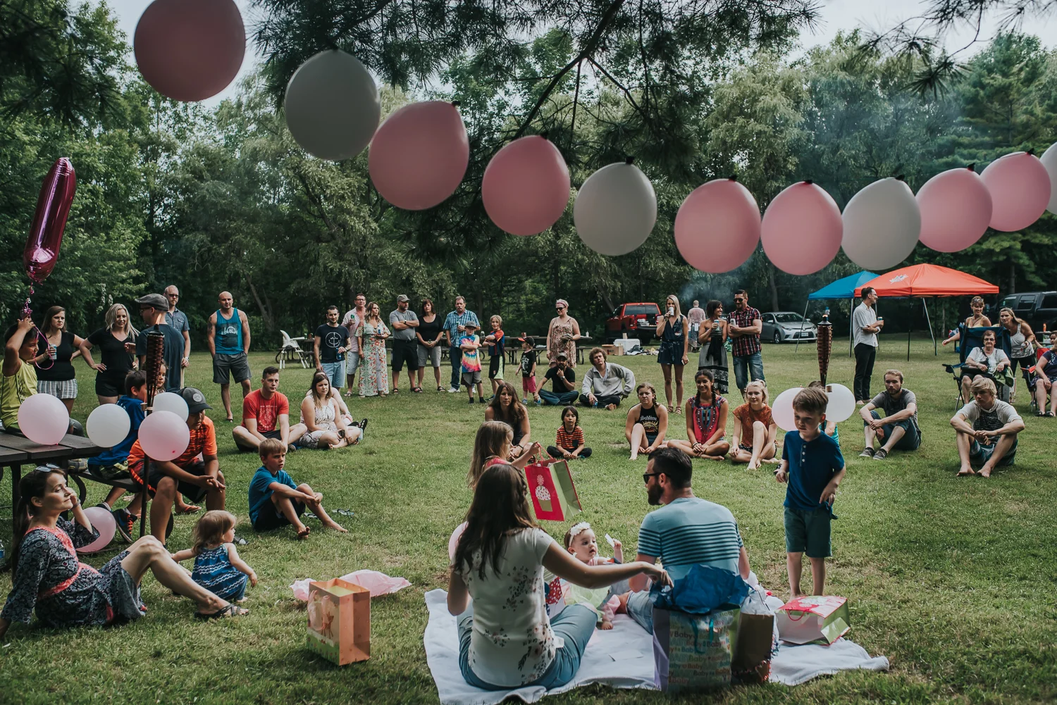 Birthday celebration with pink and white balloons gifts family sitting on a blanket surrounded by family and friends sitting and standing and watching in a camp ground wooded setting