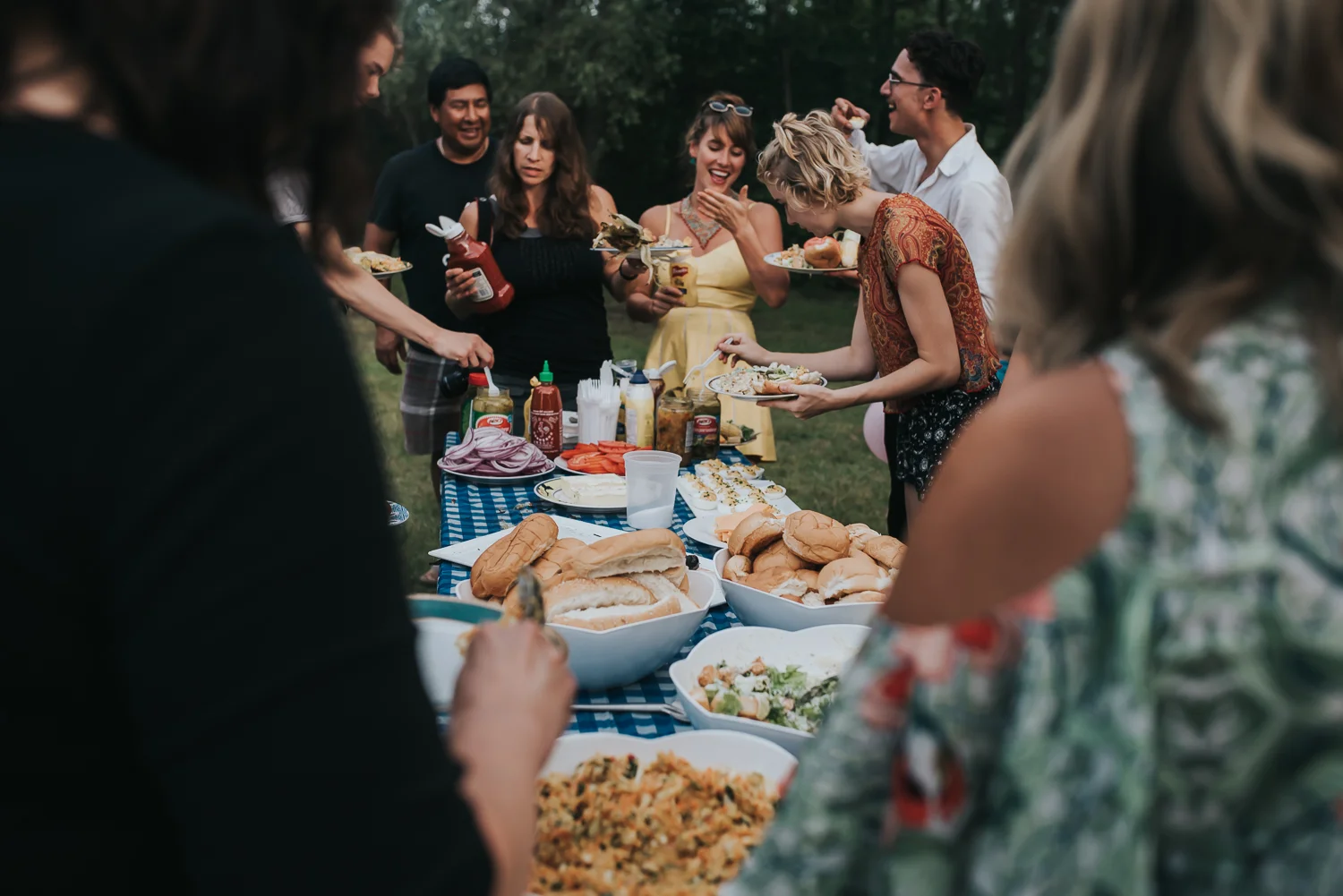 Family gathered around a BBQ meal while camping including salads, buns, hamburger and hotdogs and all the fixins.