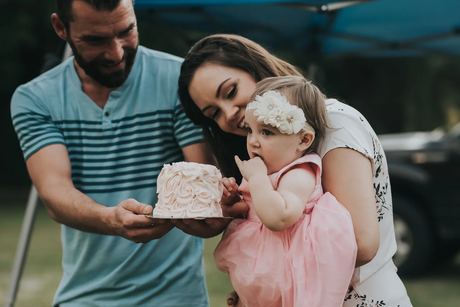 Baby in a pink dress and flowers in her hair being held by mom dressed in a white floral shirt and dad in a blue striped shirt is holding out a small blush frosted cake and baby is tasting the frosting