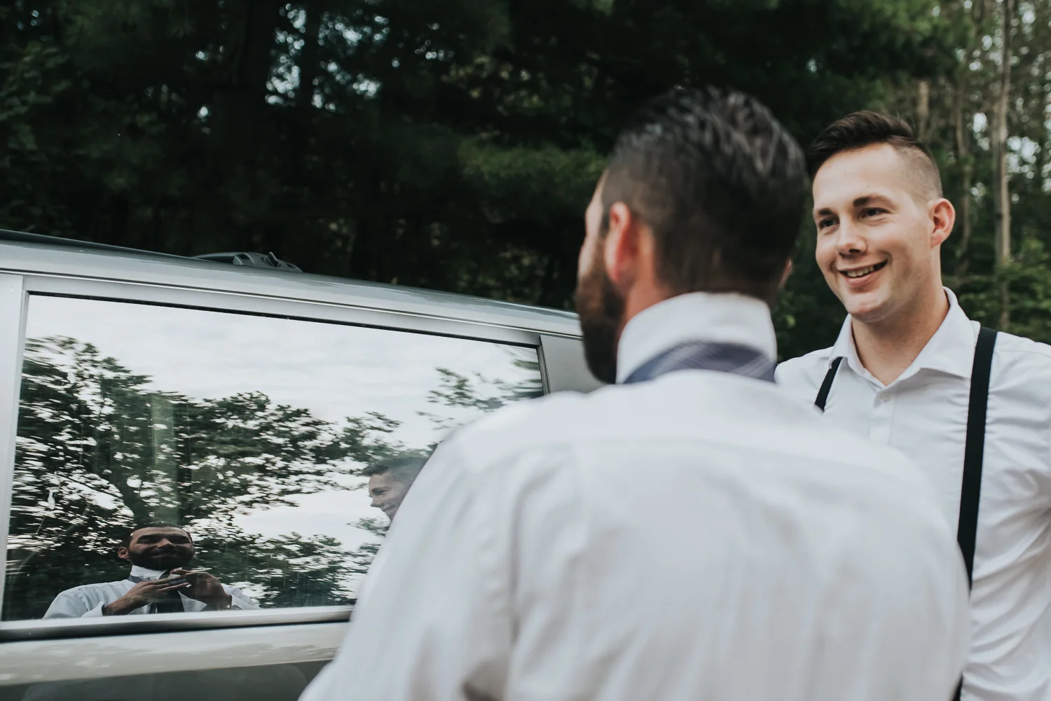 Groom putting on his tie in the reflection of a car window while his groomsman looks on both in white shirts and black suspenders