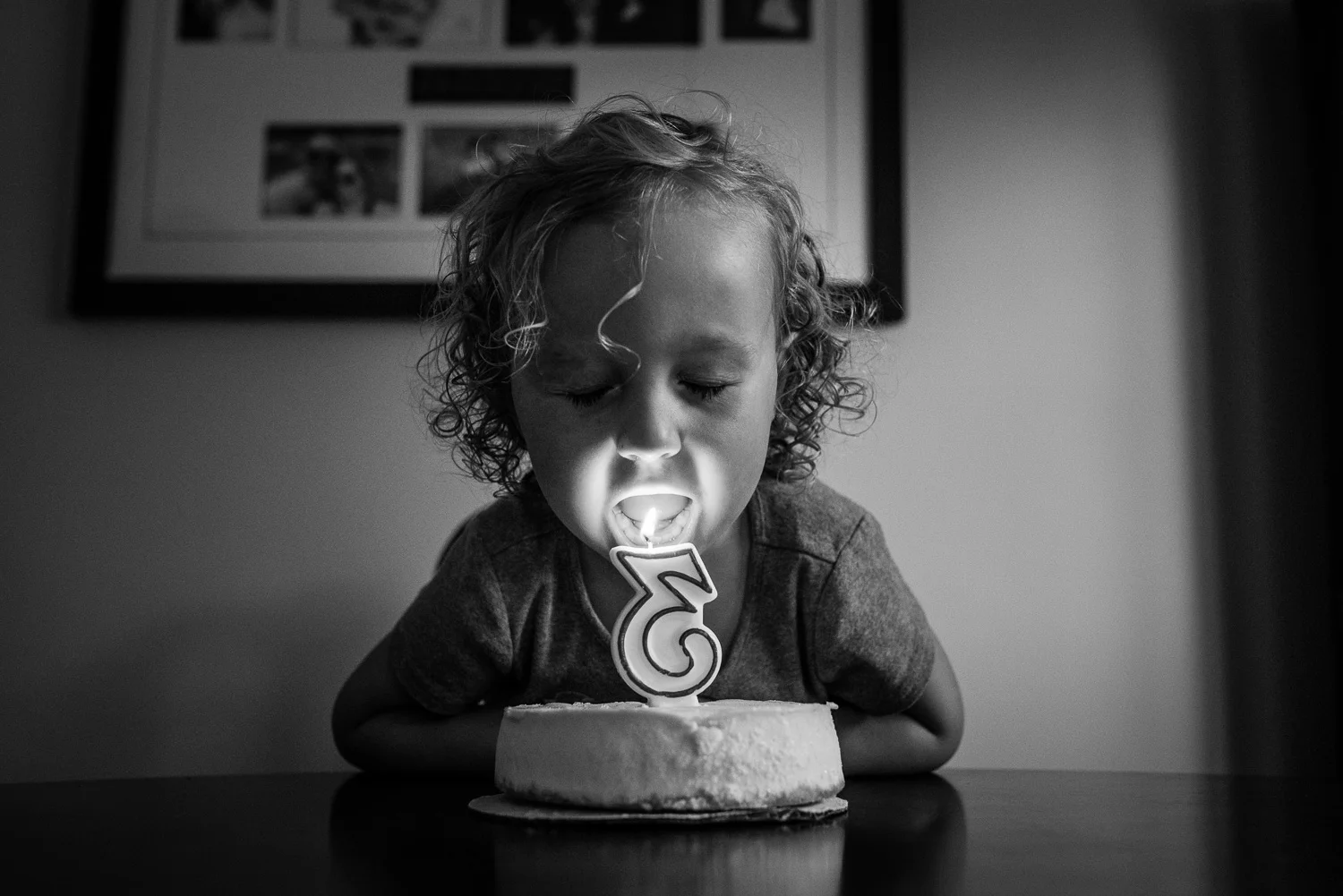 Toddler girl with curly hair at a table blowing out a number three candle on a small white cheesecake with photographs on the wall in behind her.